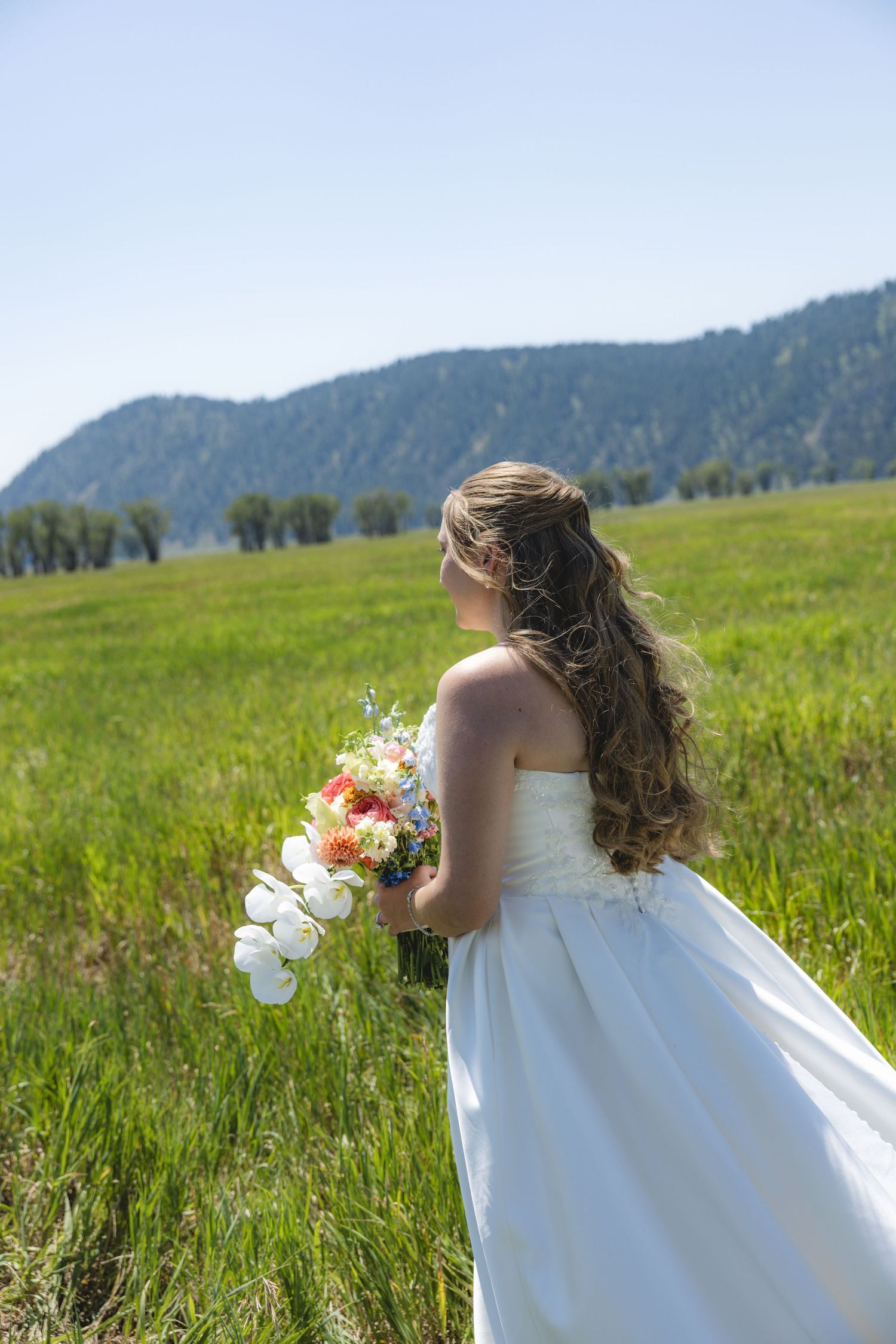 Bride in white strapless gown holding colorful bouquet stands in green meadow with mountain backdrop on sunny day.