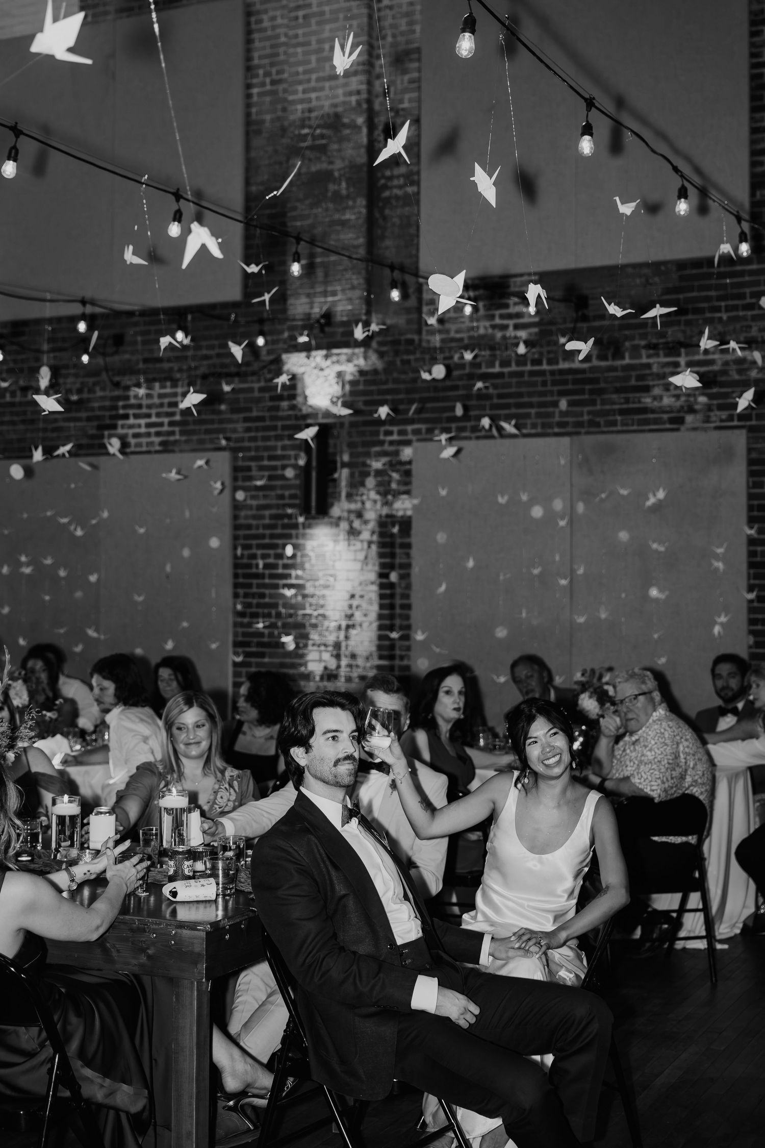 Black and white wedding reception photo with couple seated amid origami crane decorations and string lights in a brick venue.