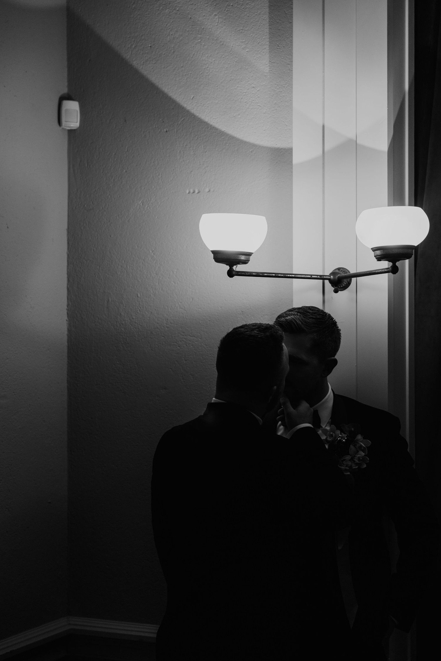 Black and white photo of two grooms sharing an intimate moment near wall sconces on their wedding day.