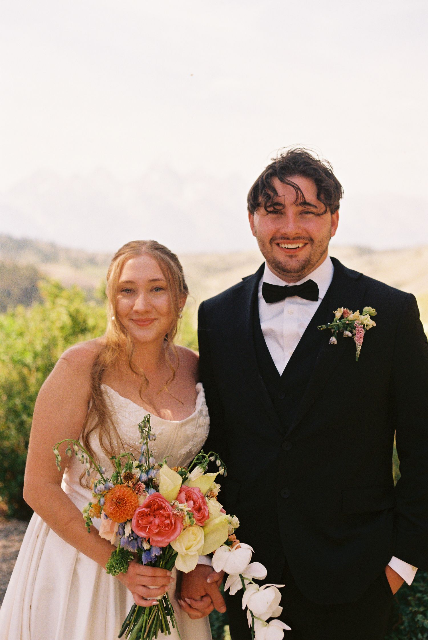 Smiling bride holding colorful bouquet stands beside groom in black tuxedo with boutonniere, scenic hills in background.