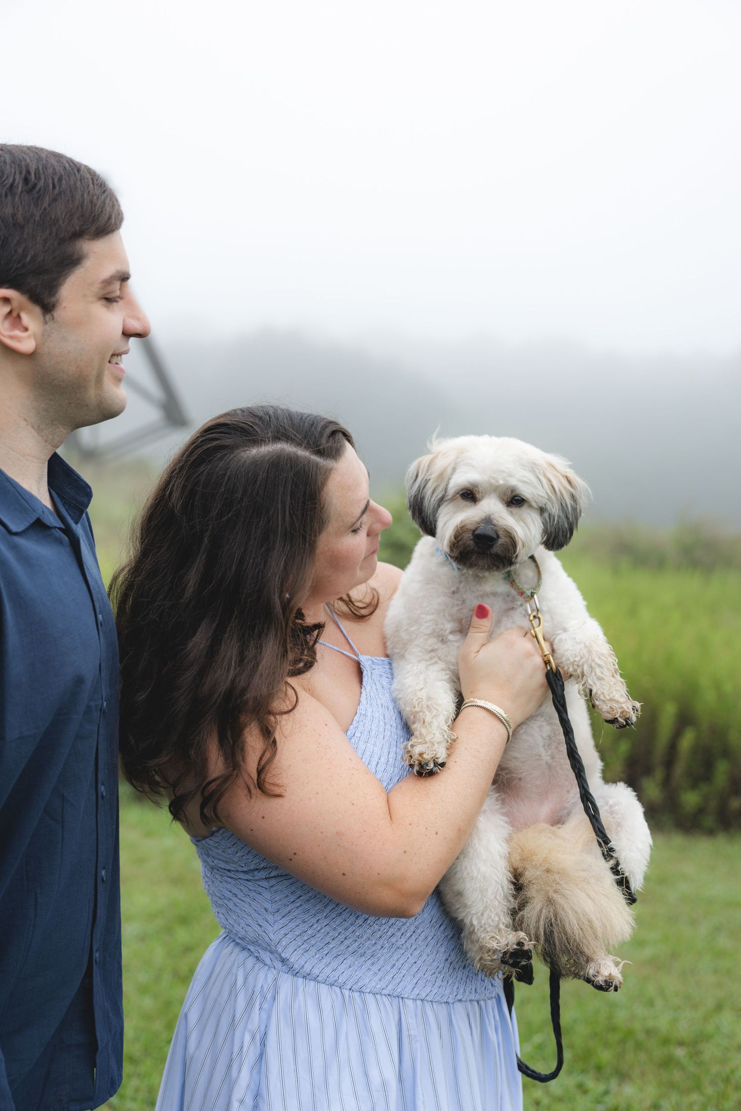 A smiling couple holds a fluffy white Shih Tzu dog outdoors on a misty green hillside, woman in blue dress.