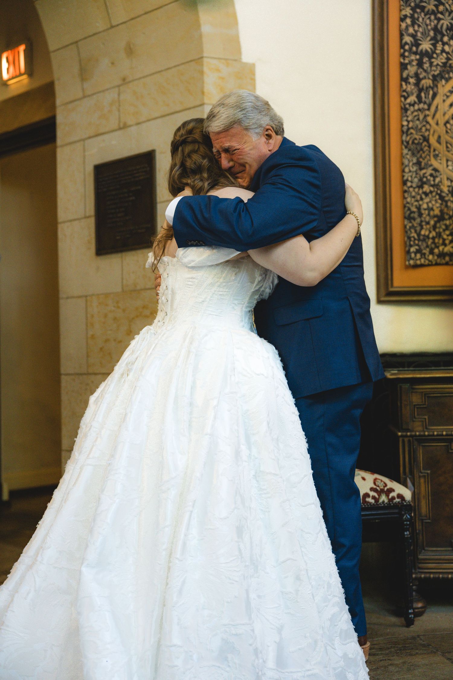 Emotional father-daughter embrace at wedding, bride in white ball gown hugging man in blue suit indoors.