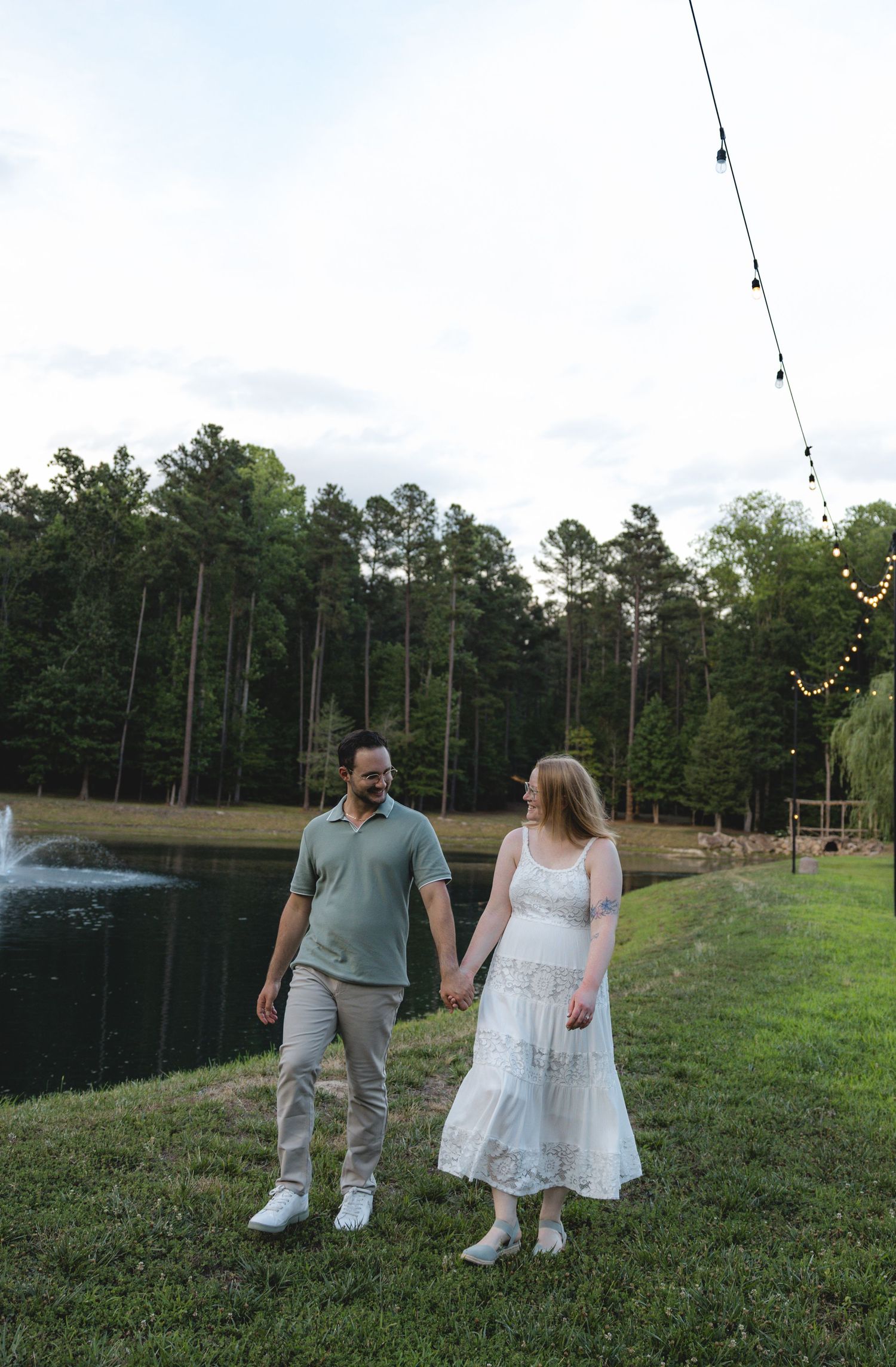 Couple holding hands walking by a pond with string lights and trees at dusk.