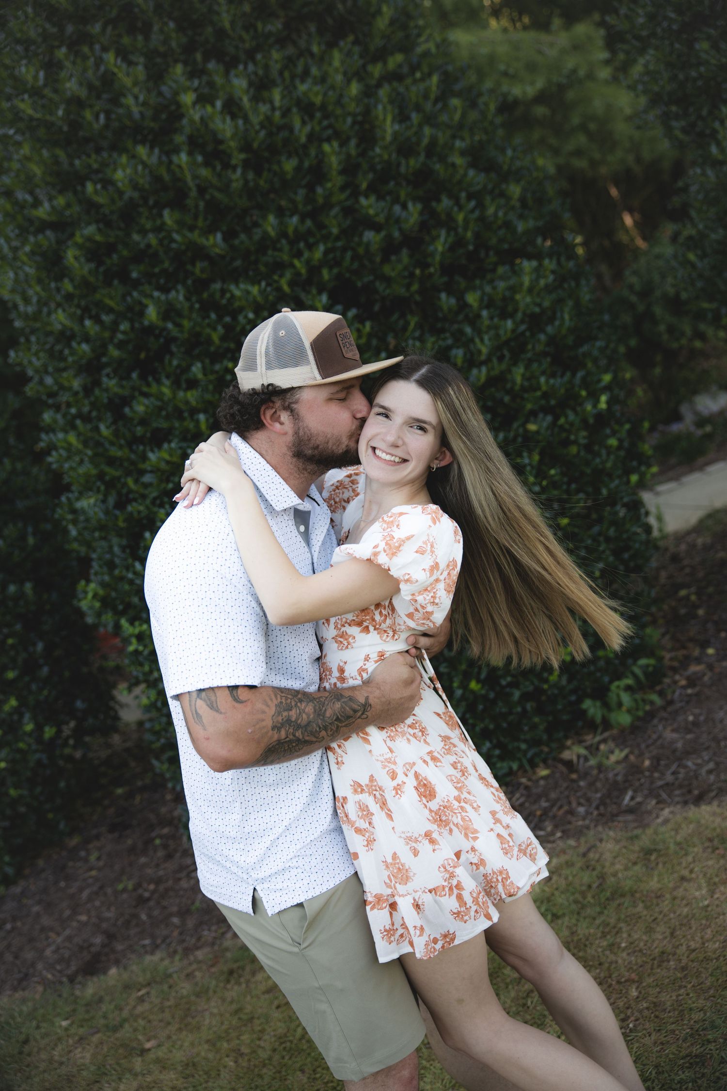 A couple embraces outdoors, man in cap kisses woman in floral dress near green bushes.