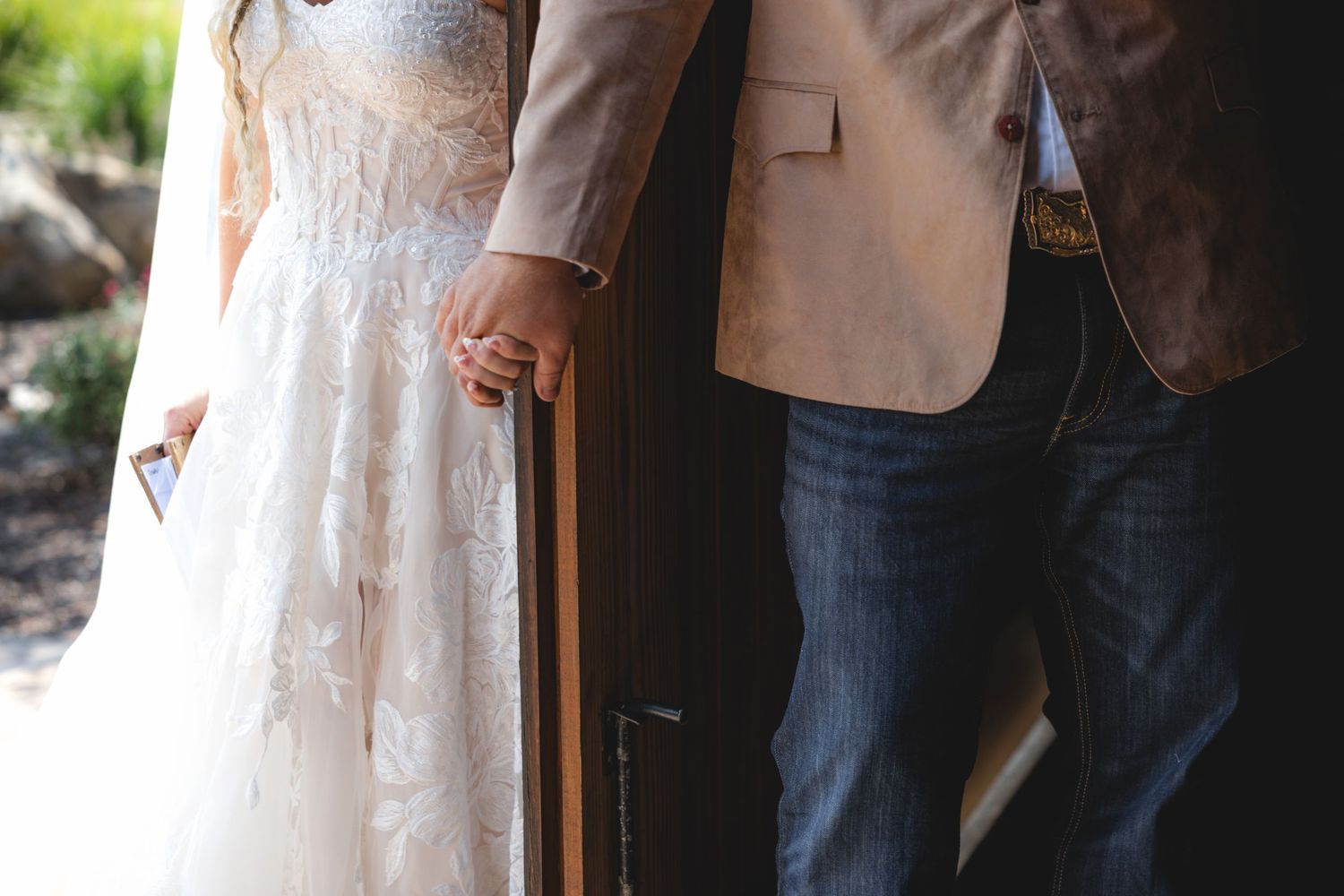 Bride and groom holding hands at wedding venue door, wearing lace gown and tan blazer with jeans.