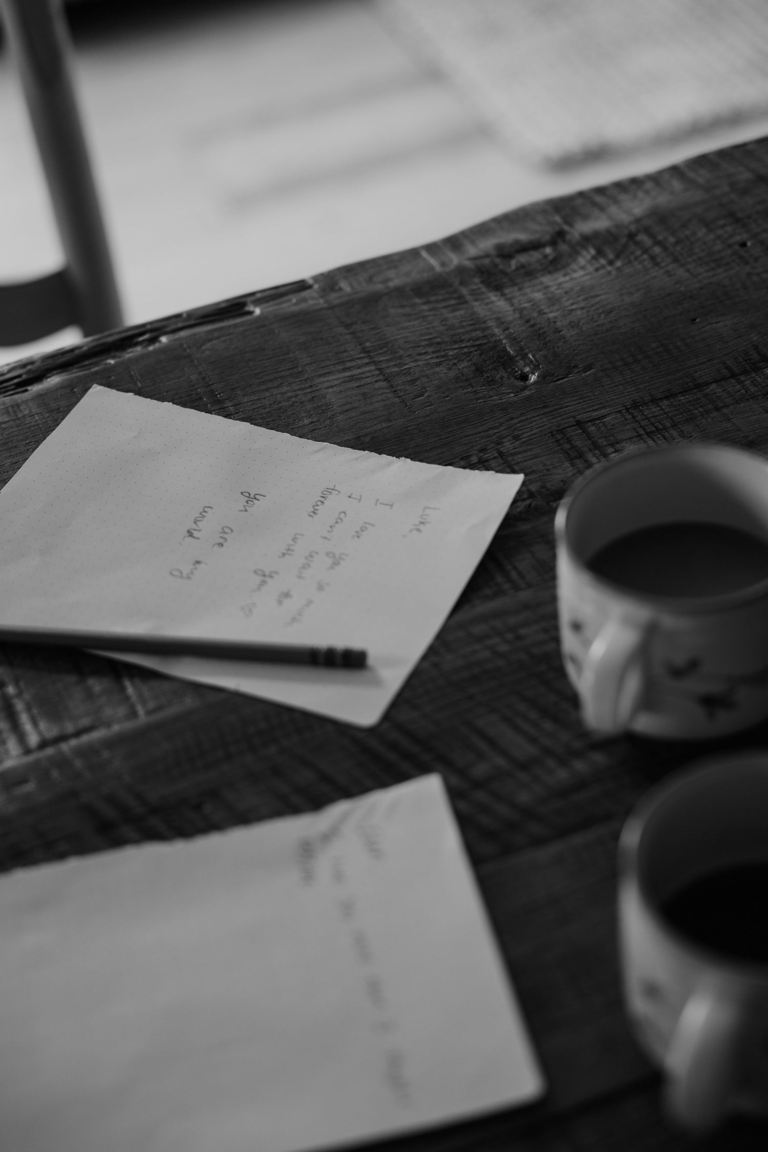 Black and white photo of handwritten notes on paper beside two coffee mugs on a rustic wooden table.