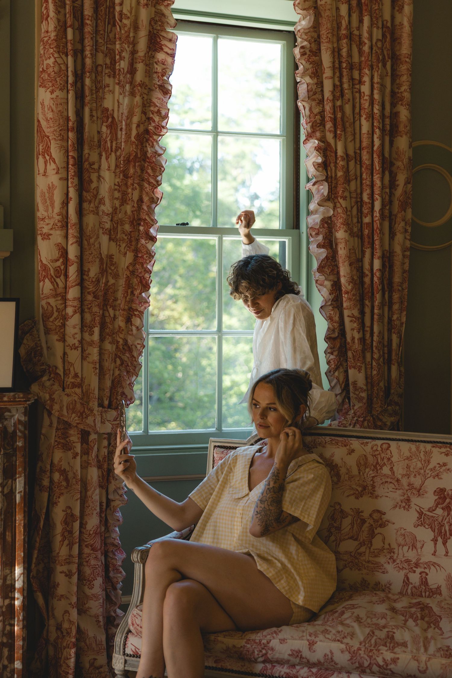 Two women pose elegantly in a vintage room with floral toile curtains and sofa near a green-framed window.