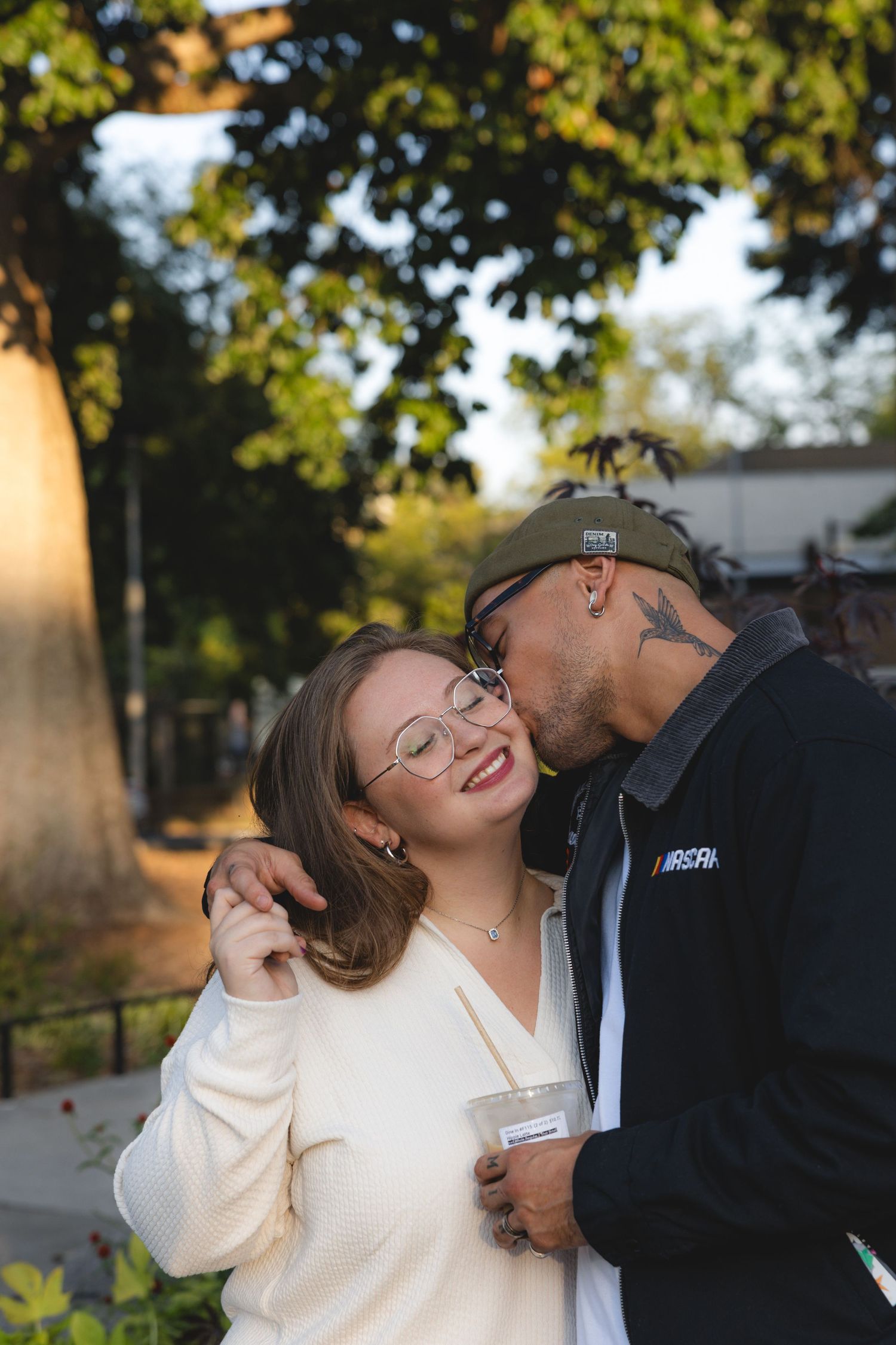 A couple shares a tender kiss outdoors, woman in white sweater holding a drink, man in black jacket, trees in background.