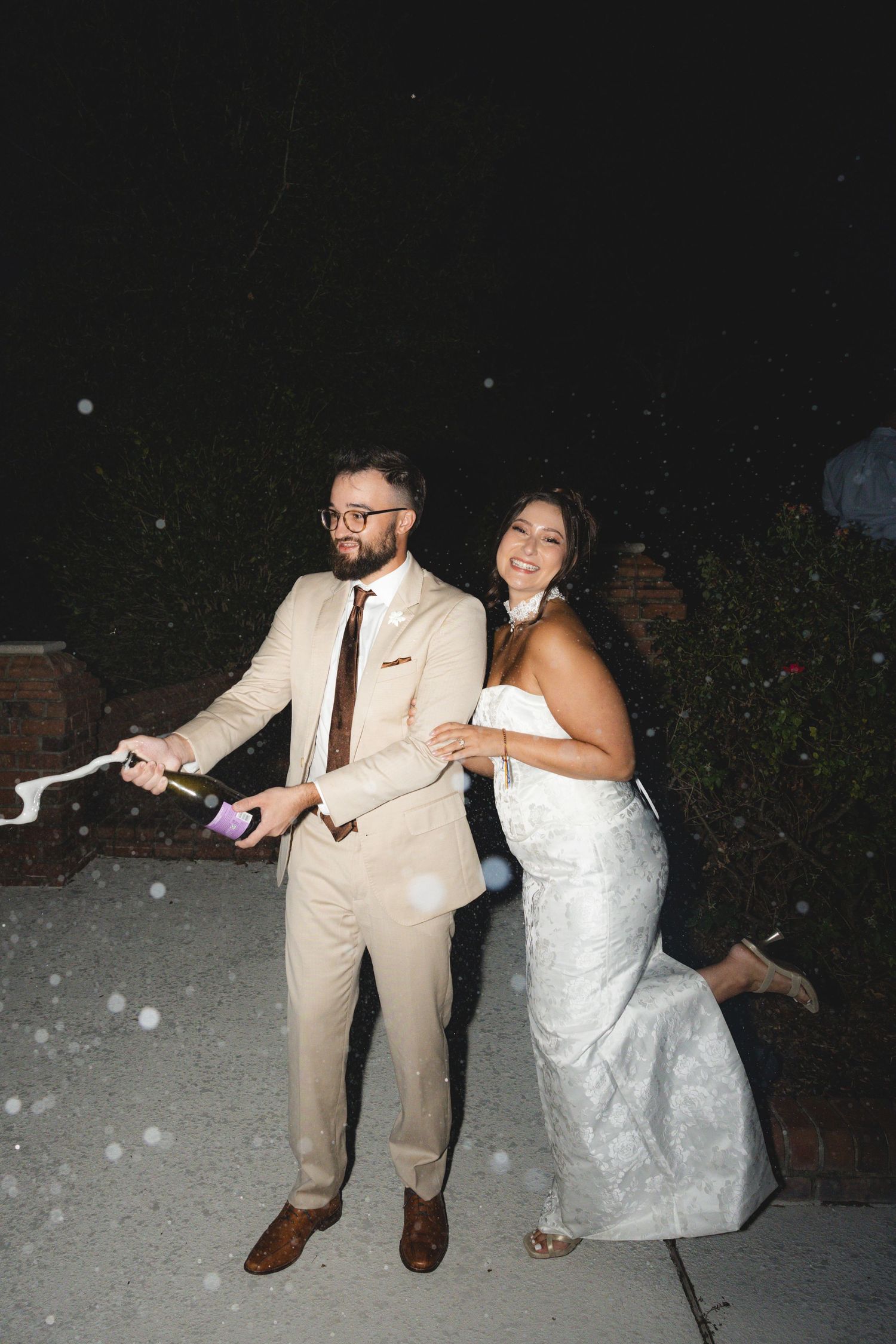 Bride and groom popping champagne bottle outdoors at night, celebrating their wedding reception.