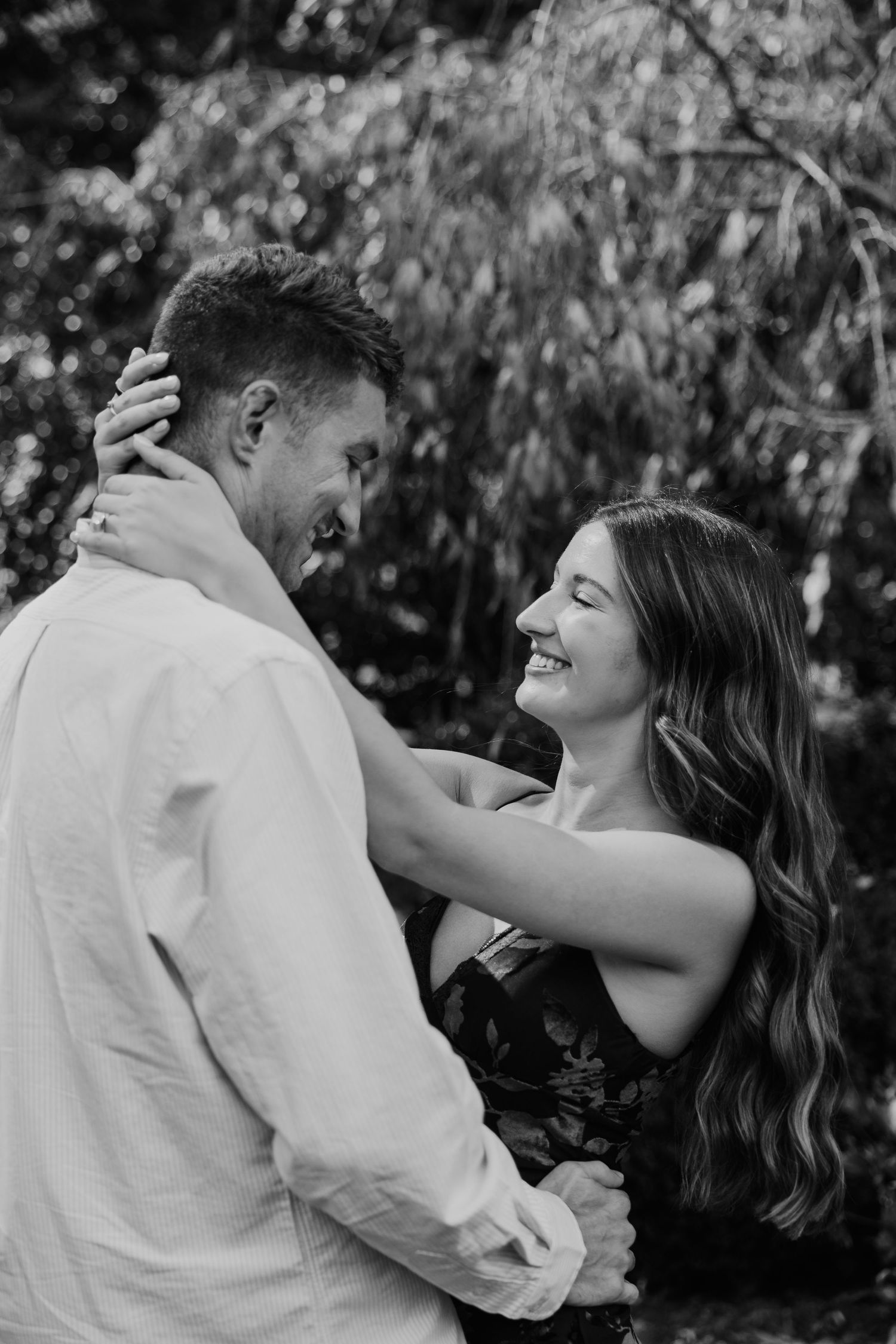 Black and white photo of a smiling couple embracing outdoors, woman laughing joyfully while holding mans face.