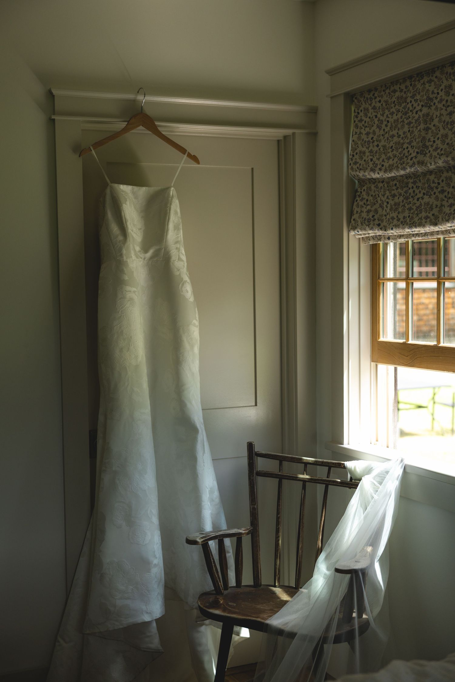Elegant white wedding dress hanging on wooden hanger near window with natural light in a cozy room with wooden chair.
