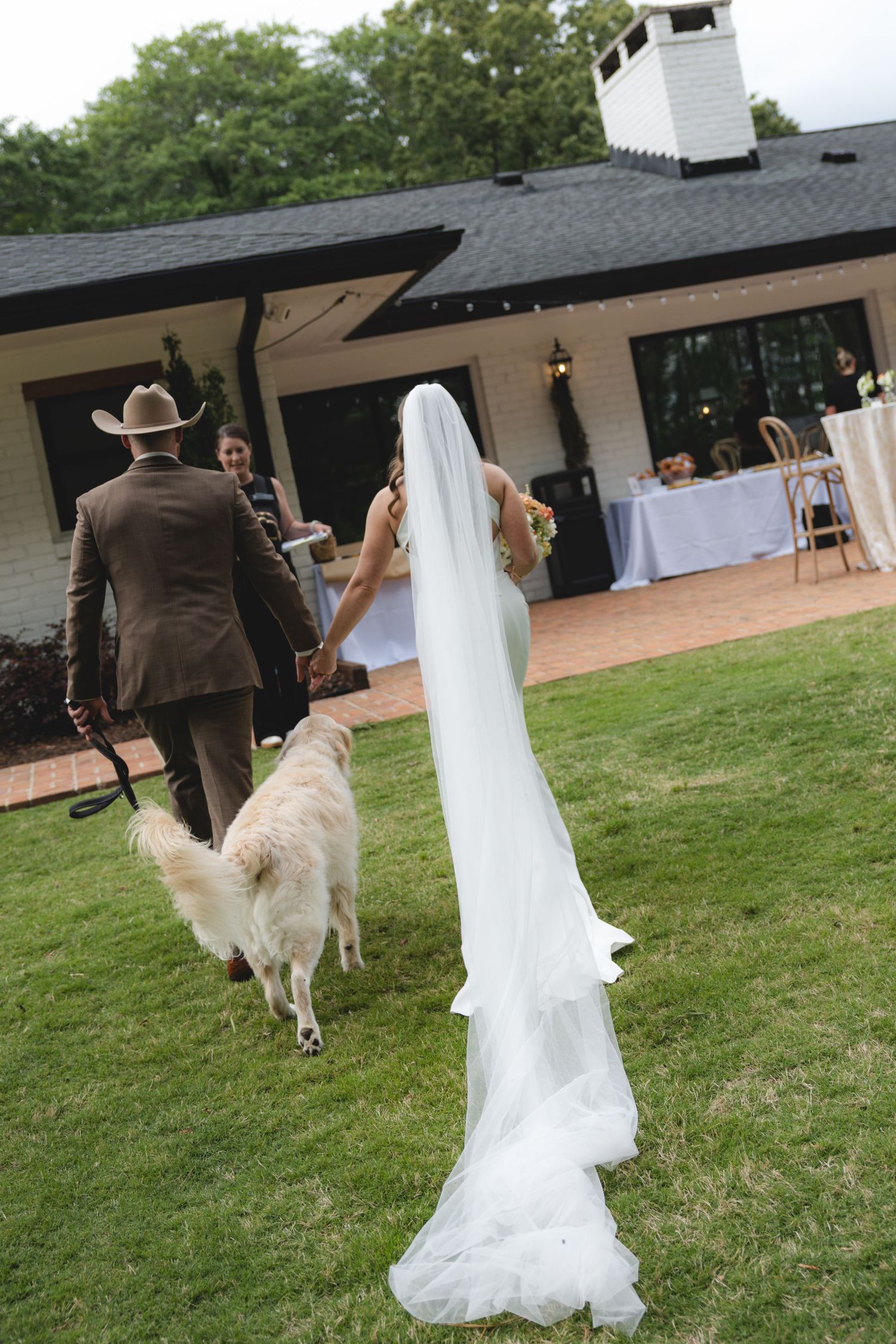 Bride in white gown and veil walks with groom in brown suit and cowboy hat alongside golden dog toward outdoor wedding reception.