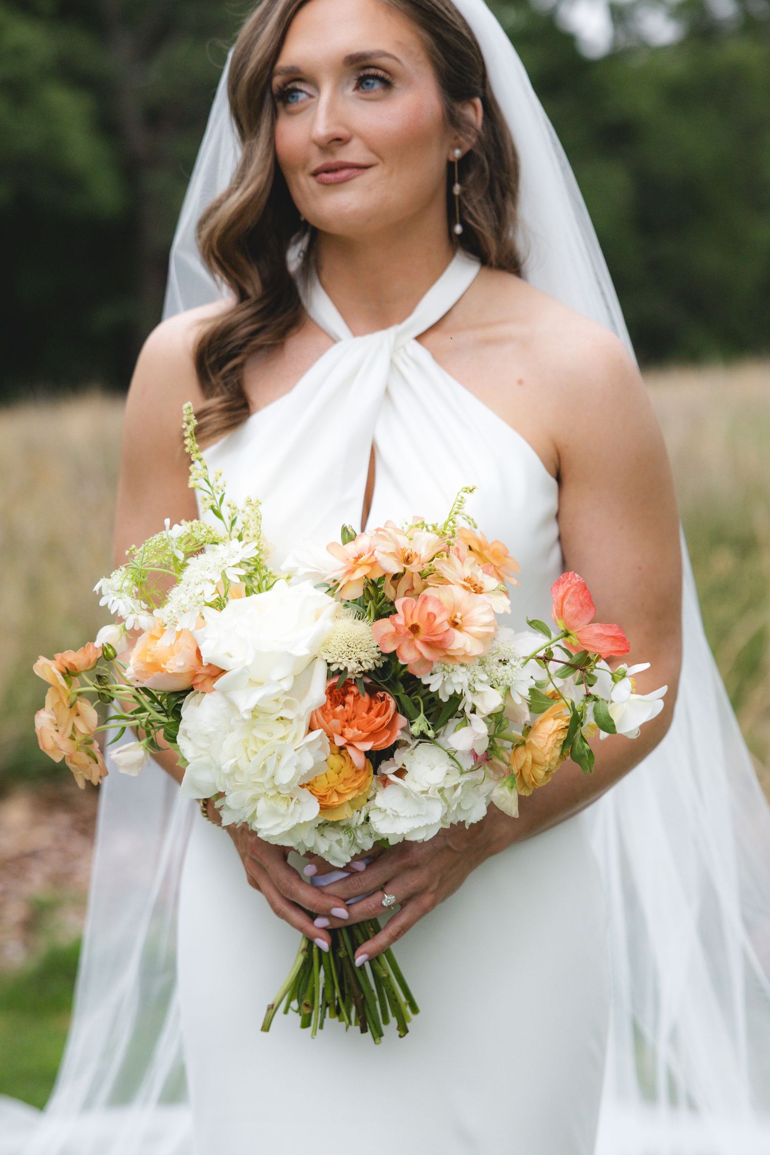 Bride in white halter gown holds peach and white floral bouquet while gazing upward wearing a veil outdoors.