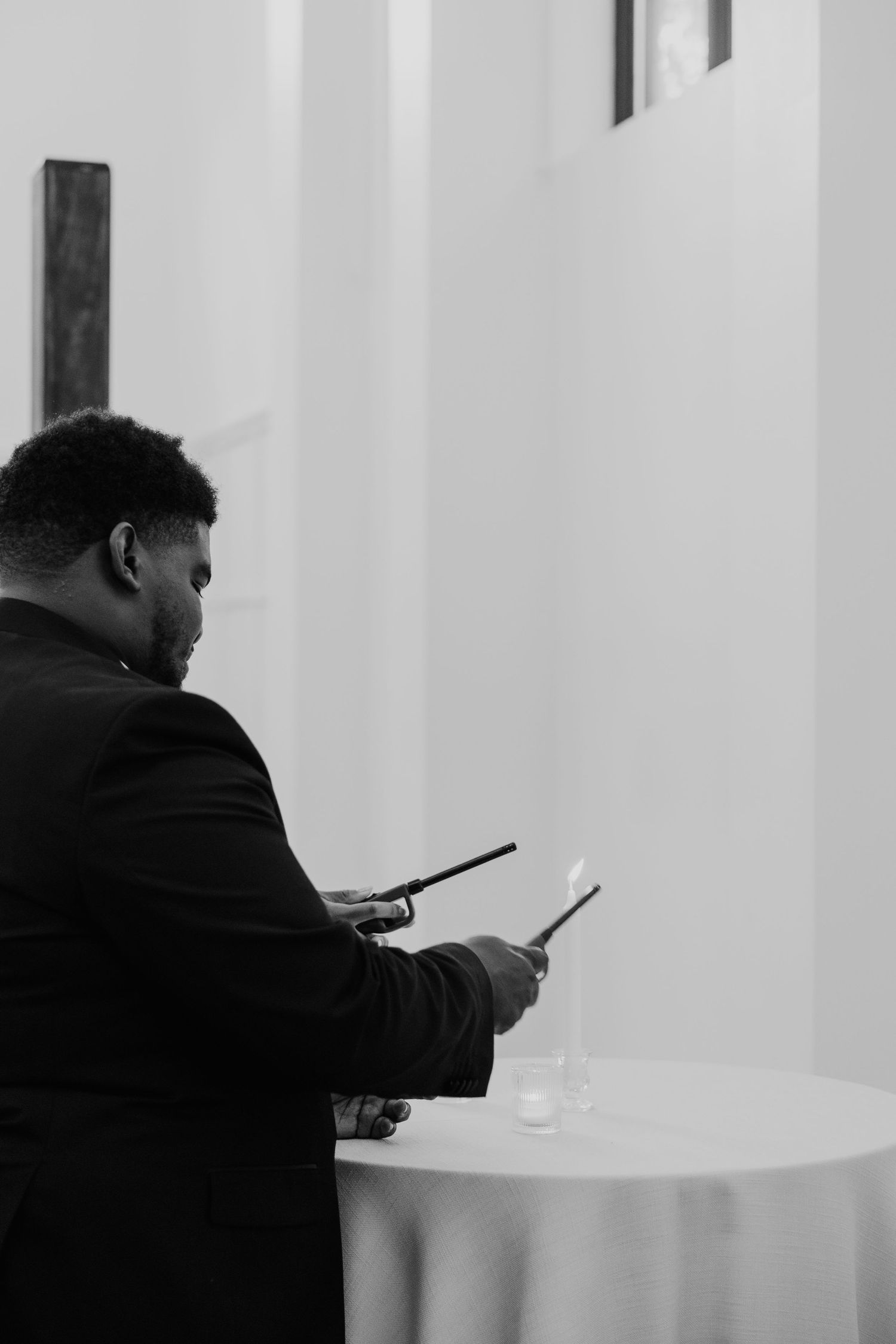 Black and white photo of groom in suit lighting candles at draped table during wedding ceremony.