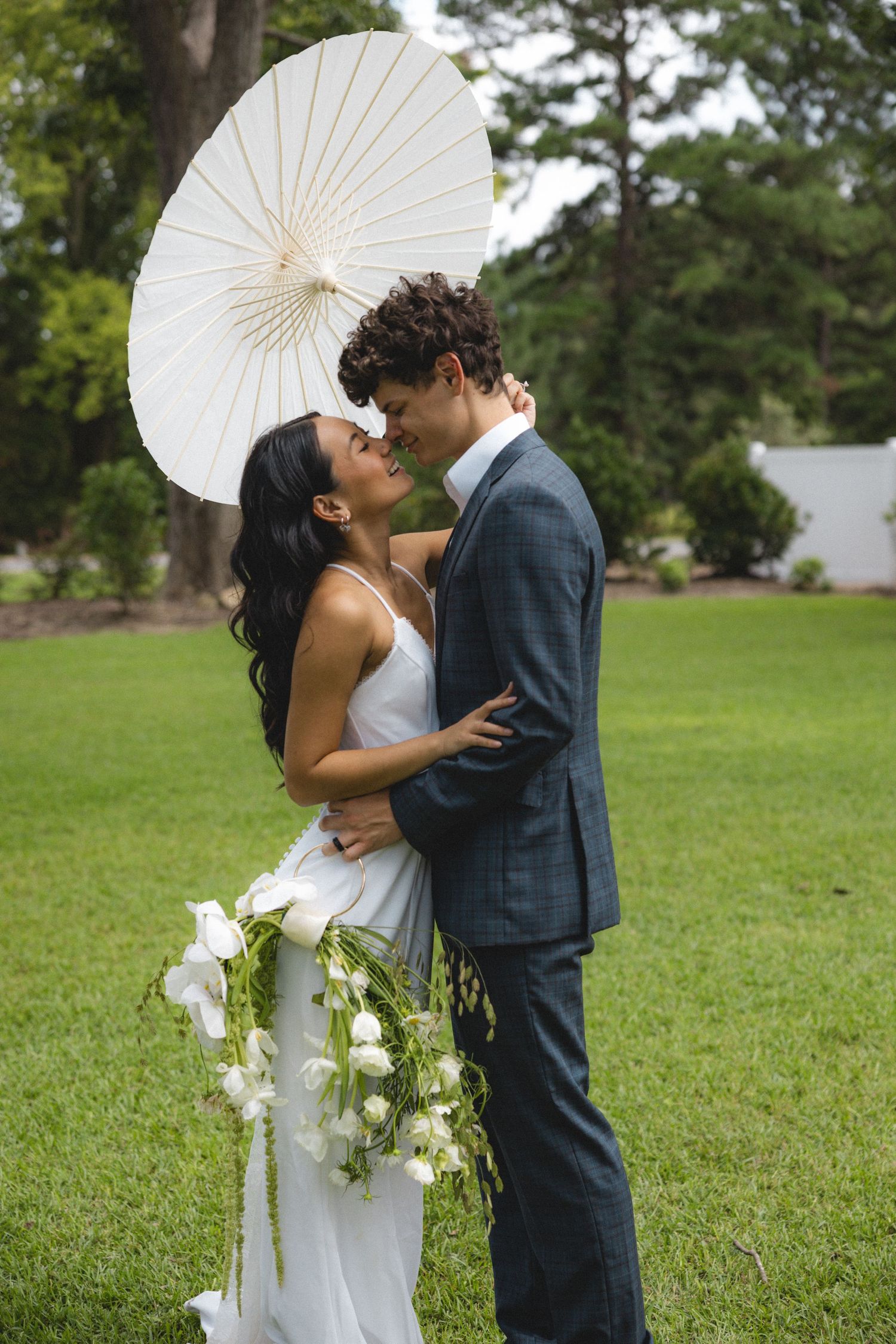 Romantic wedding couple sharing a moment under a white parasol on a green lawn.
