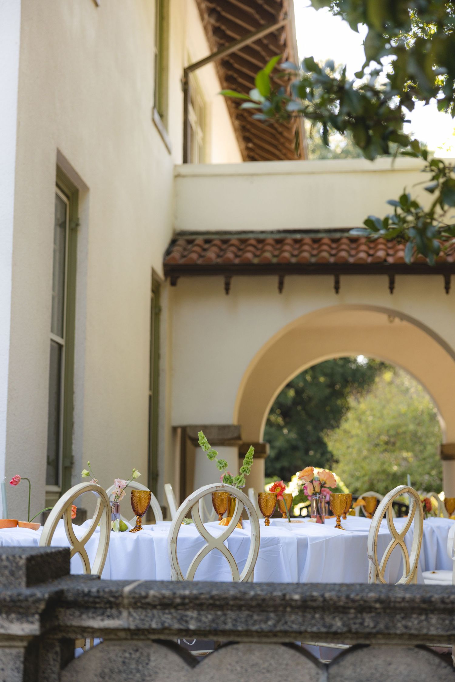 Elegant outdoor wedding reception table with white linens, amber glasses, and floral centerpieces beside a Spanish-style building.