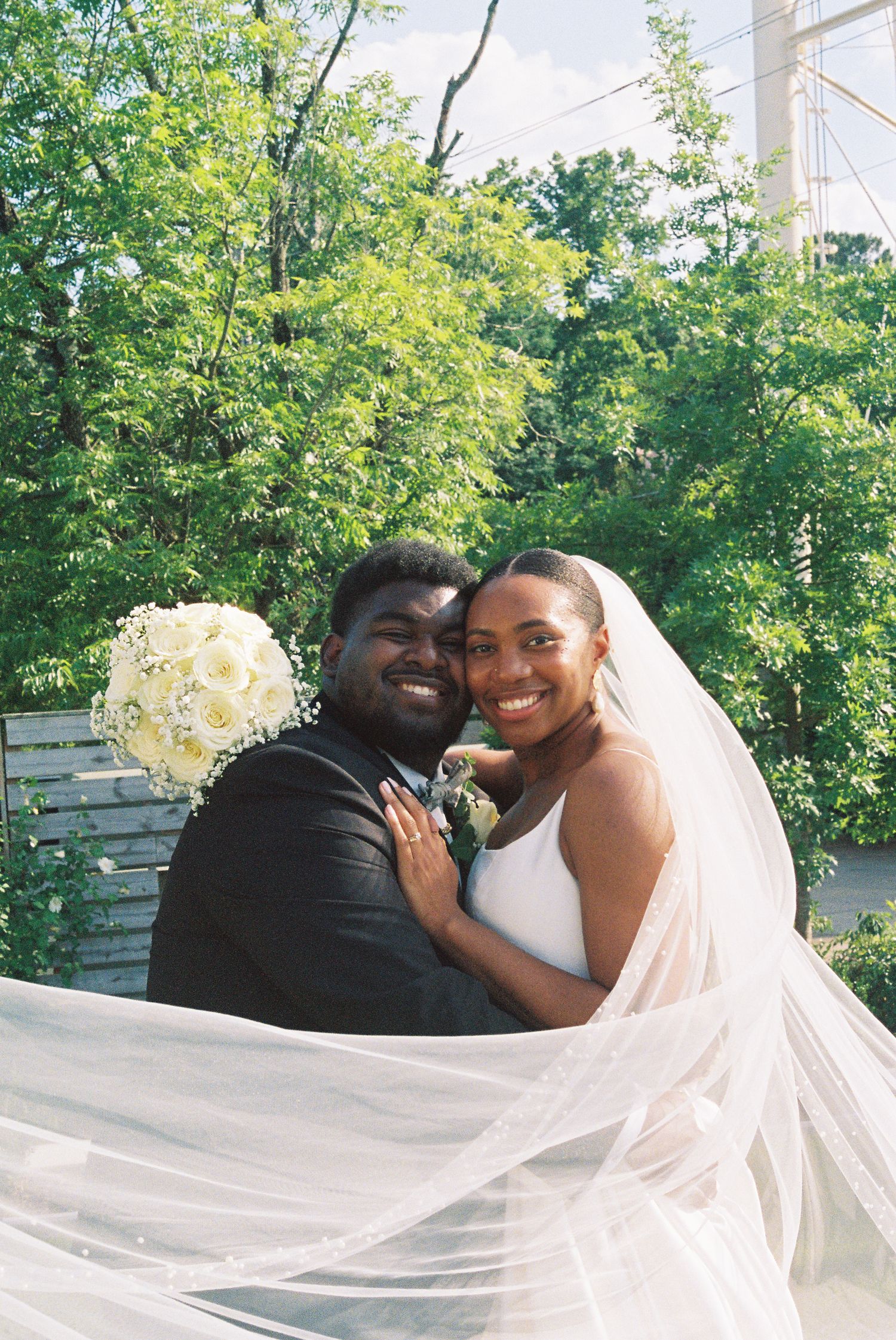 A joyful newlywed couple poses outdoors, bride in white gown with veil and groom in suit, surrounded by lush green trees.