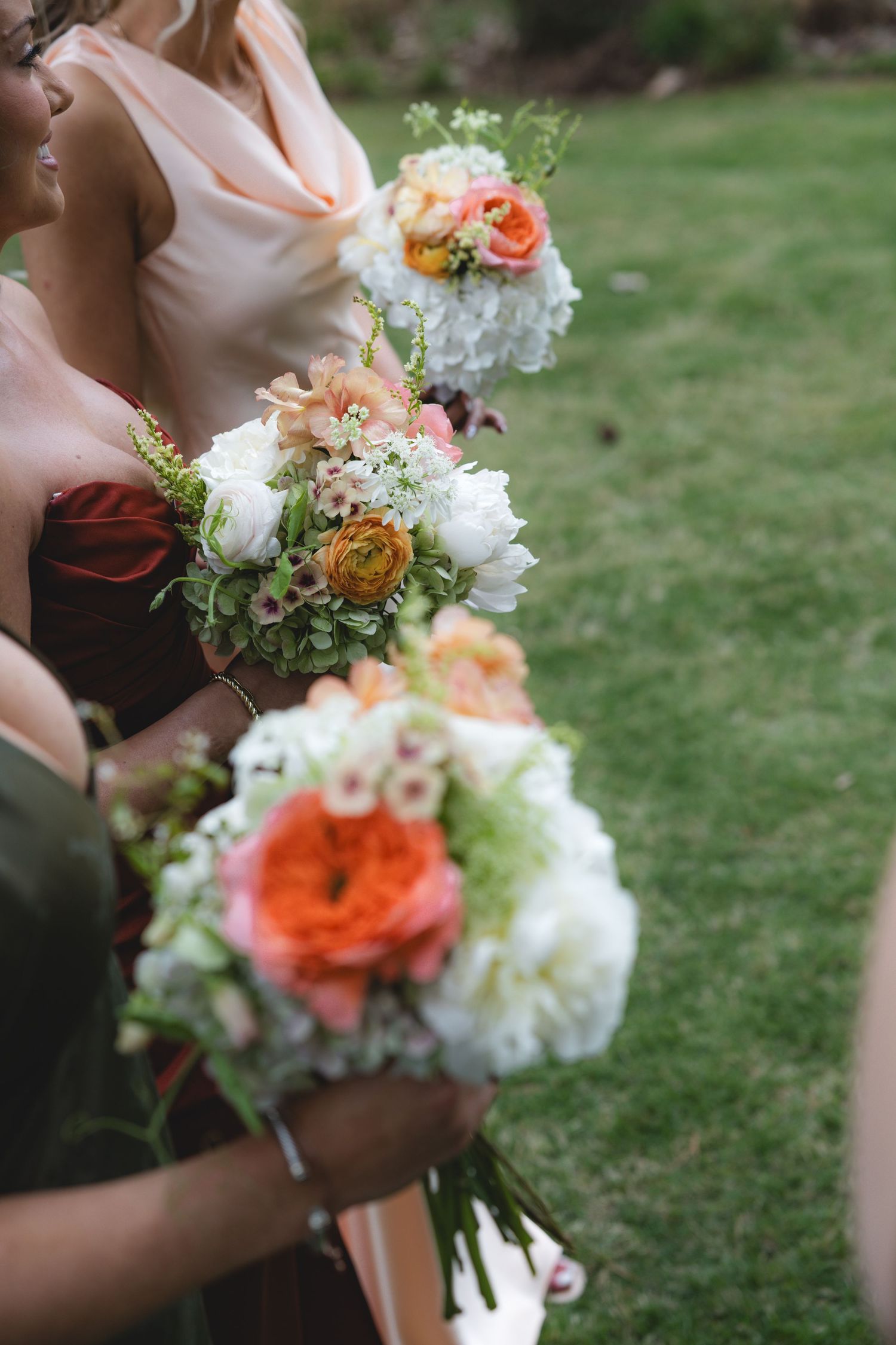 Bridesmaids hold lush bouquets of white, peach, and orange flowers including ranunculus and peonies.