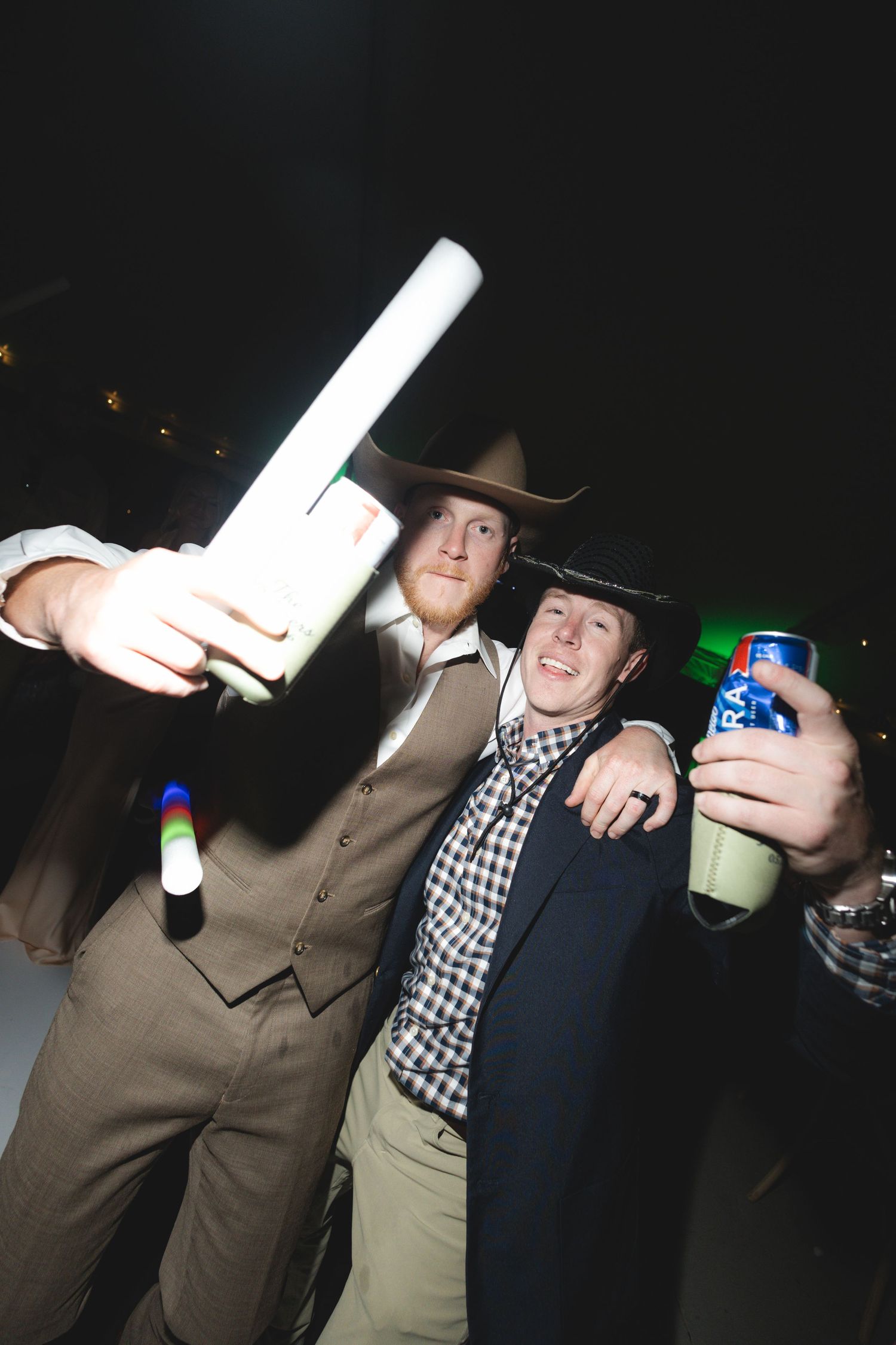 Two men in cowboy hats celebrating with drinks and glow sticks at a party.