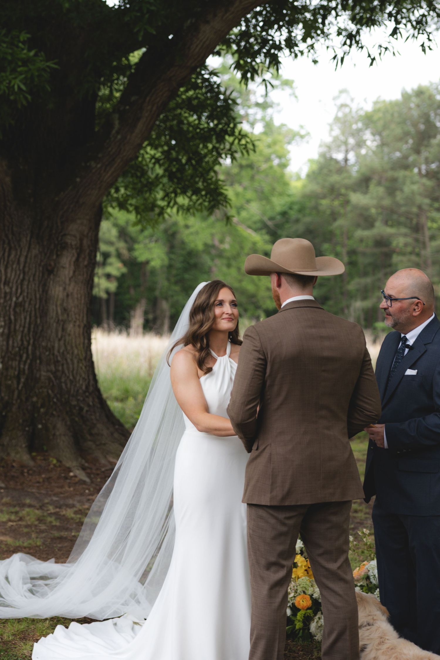 Bride in white halter gown with cathedral veil holds grooms hands during outdoor ceremony under large oak tree.