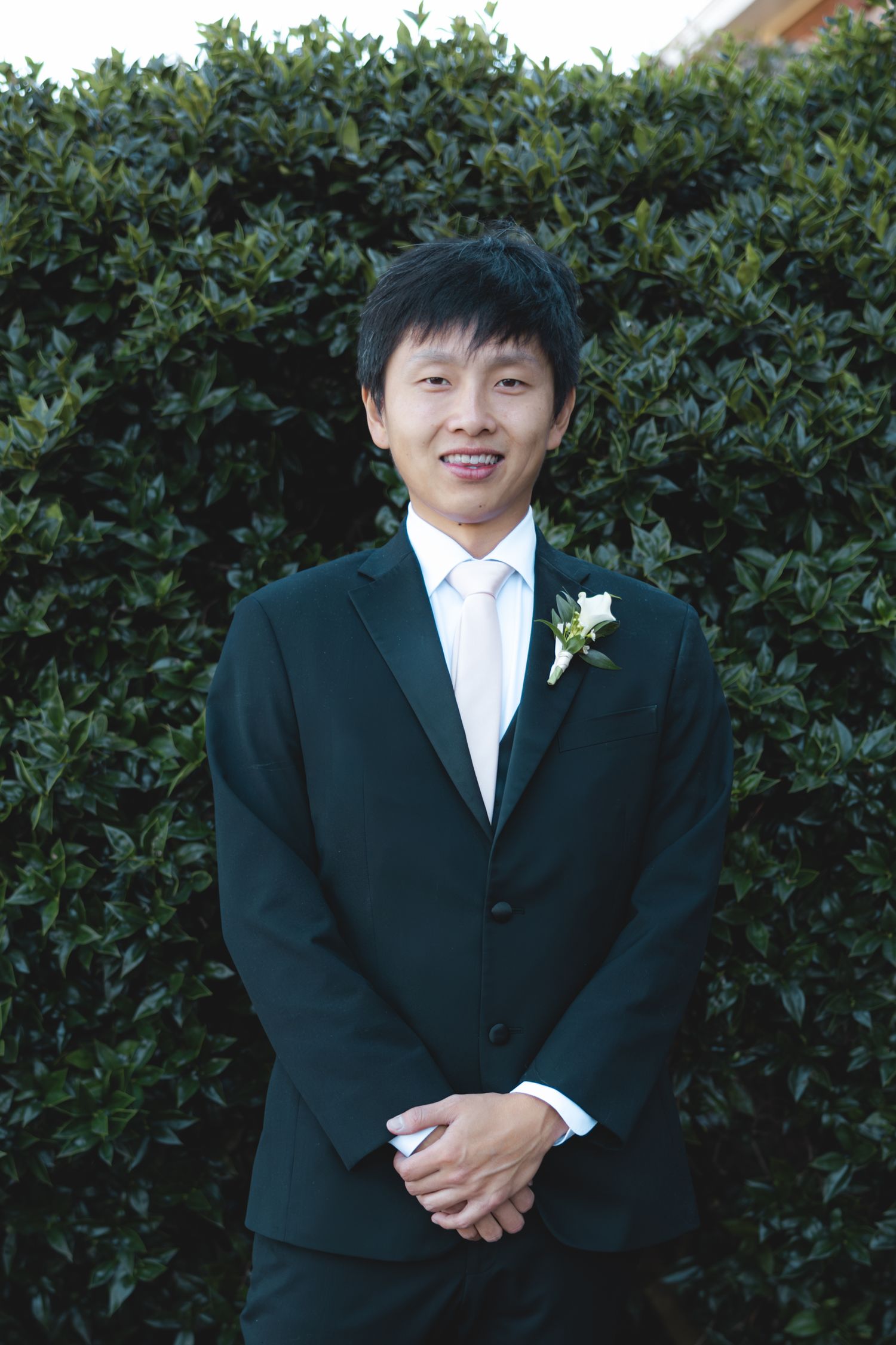 Smiling groom in black suit with white boutonniere posing in front of green hedge.