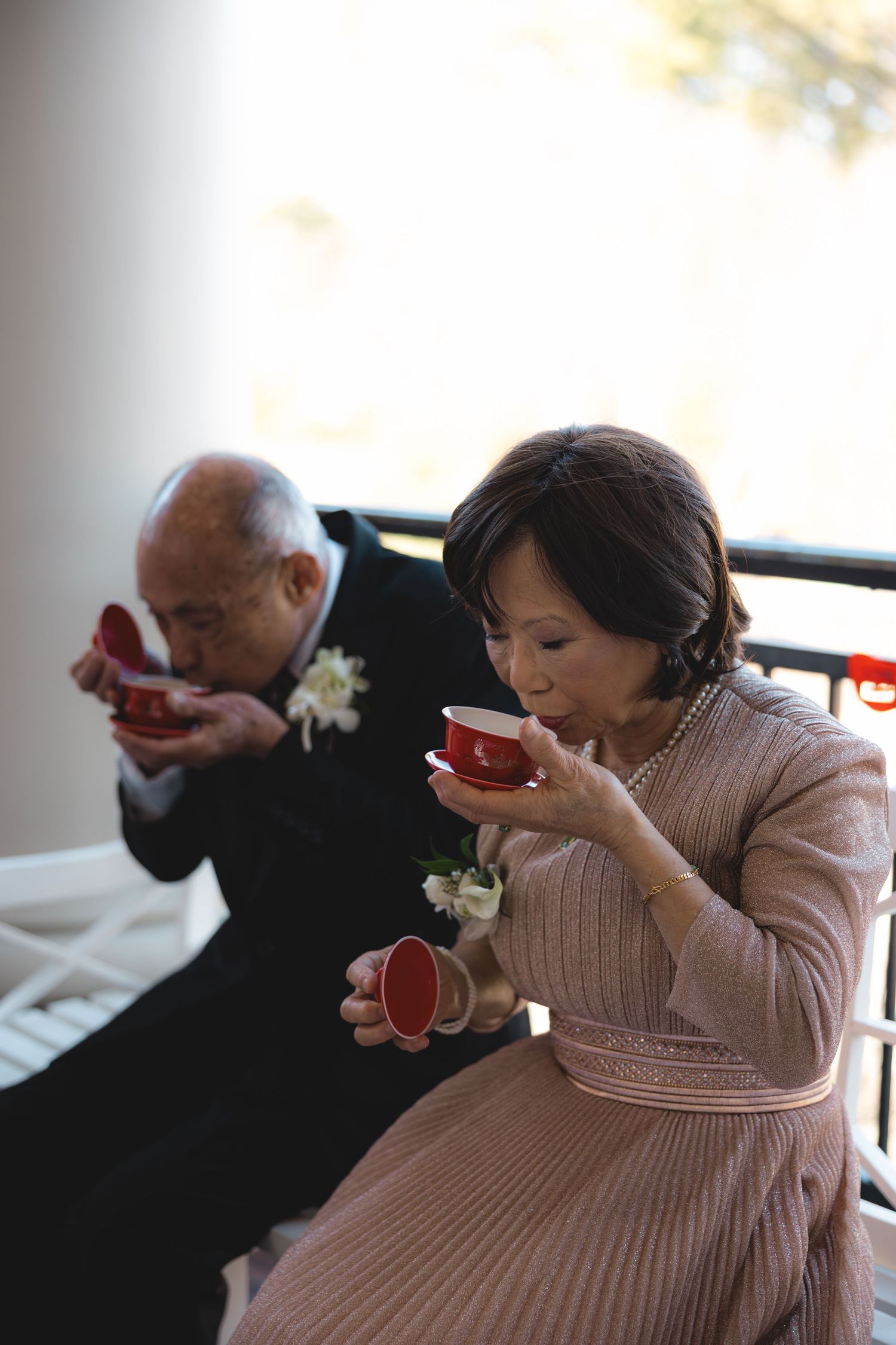 Elderly Asian couple sipping from traditional red Chinese tea cups during wedding tea ceremony.