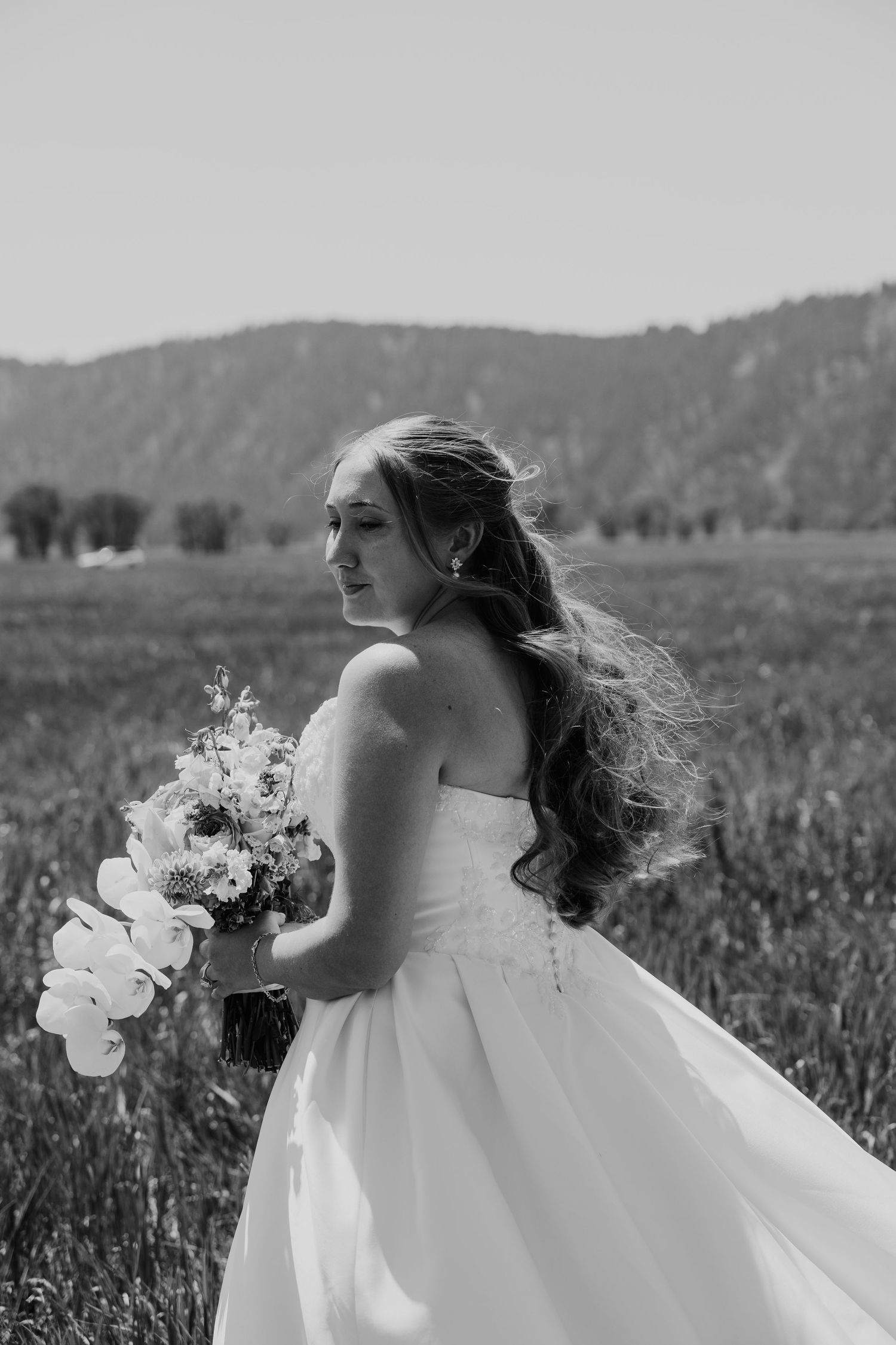 Black and white photo of a bride in a strapless gown holding a bouquet in a field with mountains behind her.