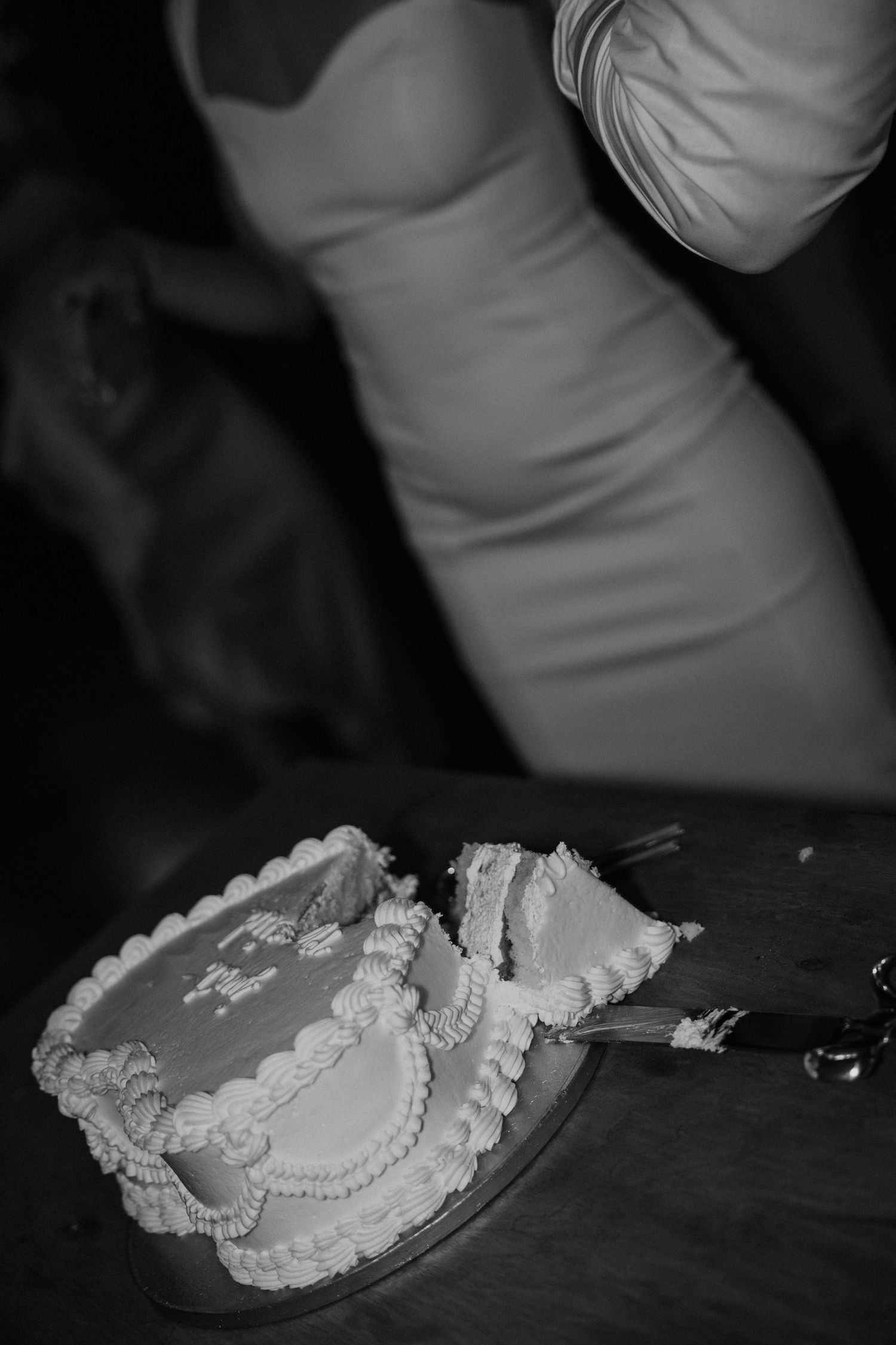 Black and white photo of a sliced wedding cake on a table with a bride in a white dress standing nearby.