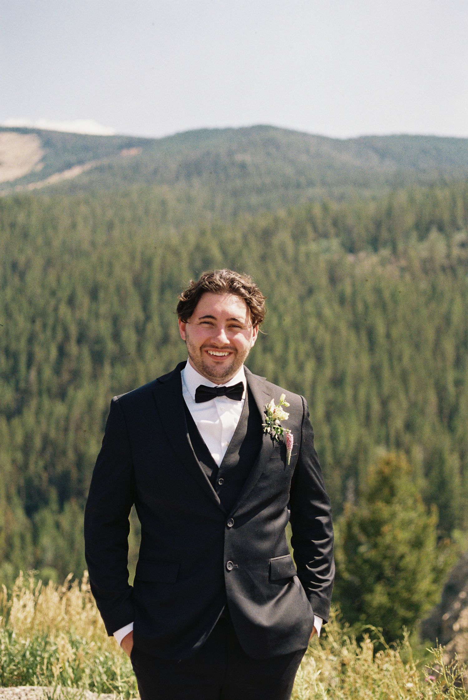 Smiling groom in black tuxedo with boutonniere poses outdoors with mountain forest backdrop on wedding day.