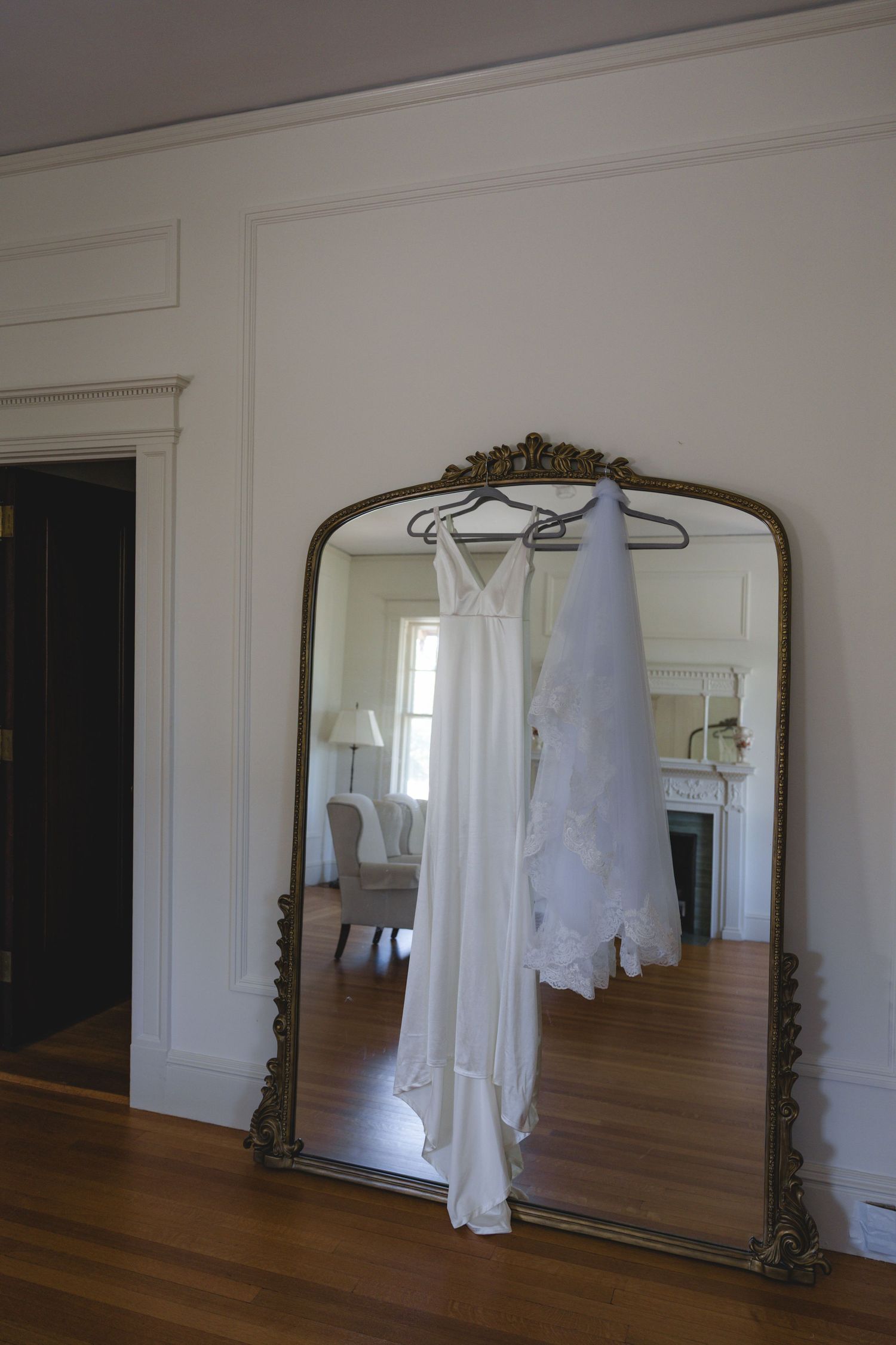 White wedding dress hanging on an ornate gold mirror in an elegant room with hardwood floors.