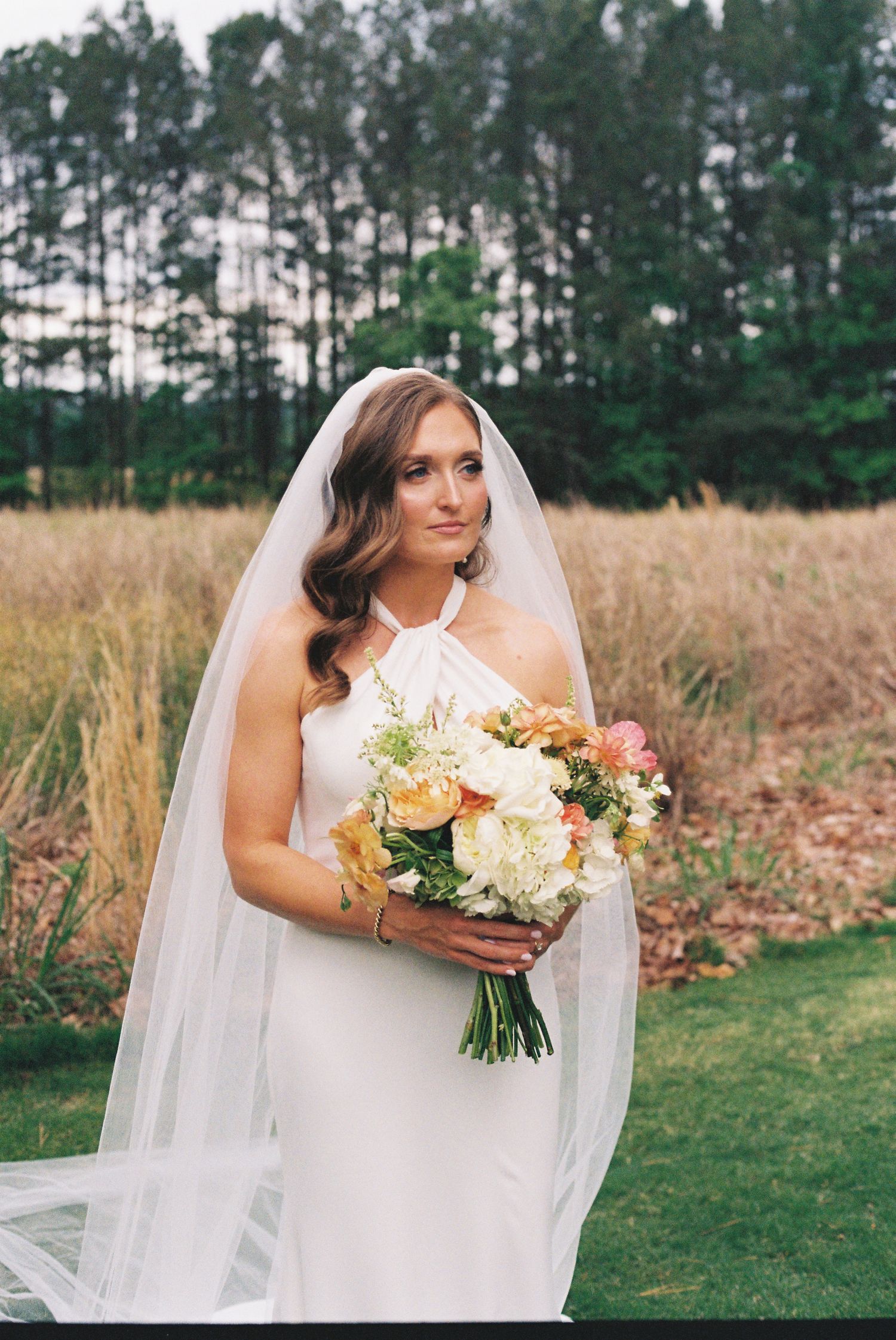 Bride in white halter gown and veil holding colorful bouquet of flowers outdoors with golden grass and trees.