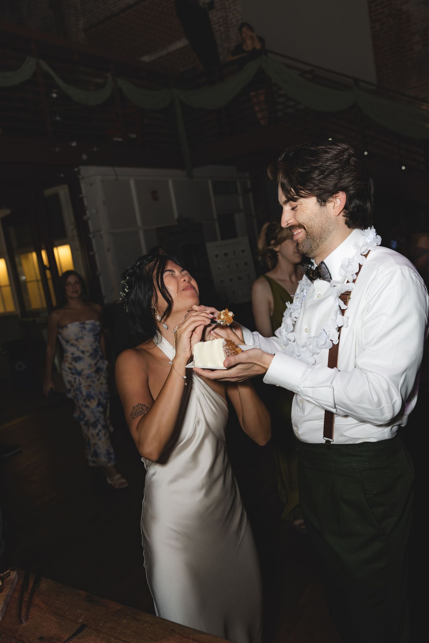 Bride and groom sharing wedding cake during reception, groom smiling in suspenders and bow tie.