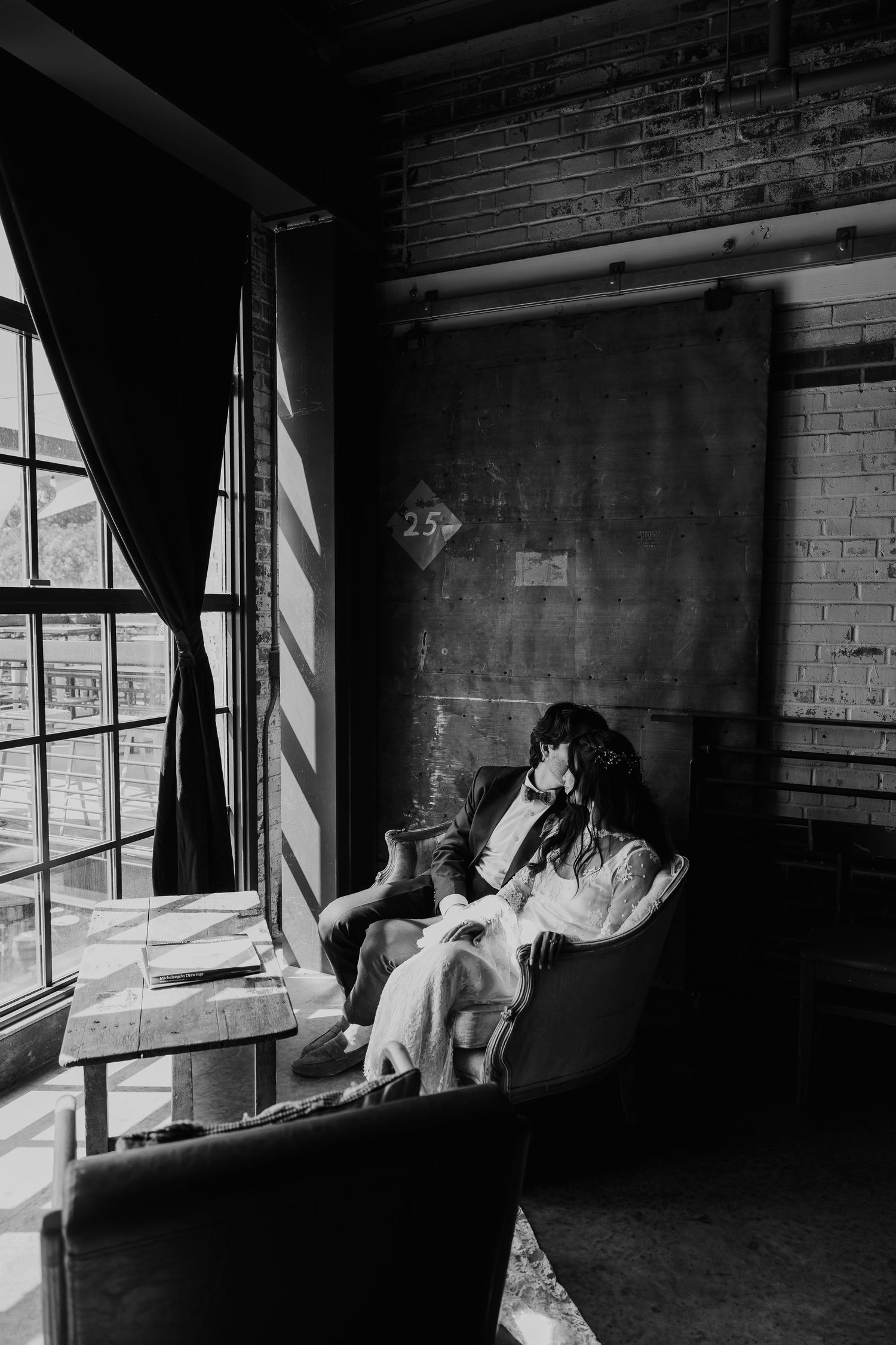 Black and white photo of a couple embracing in an armchair by industrial windows in a loft space.