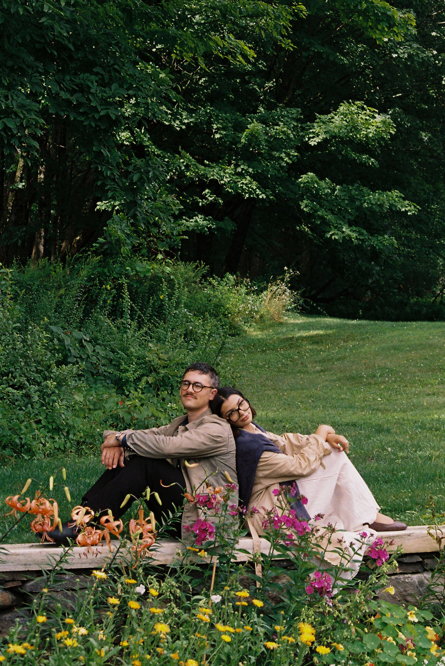 Couple sitting back-to-back on a stone wall surrounded by wildflowers in a lush green garden.