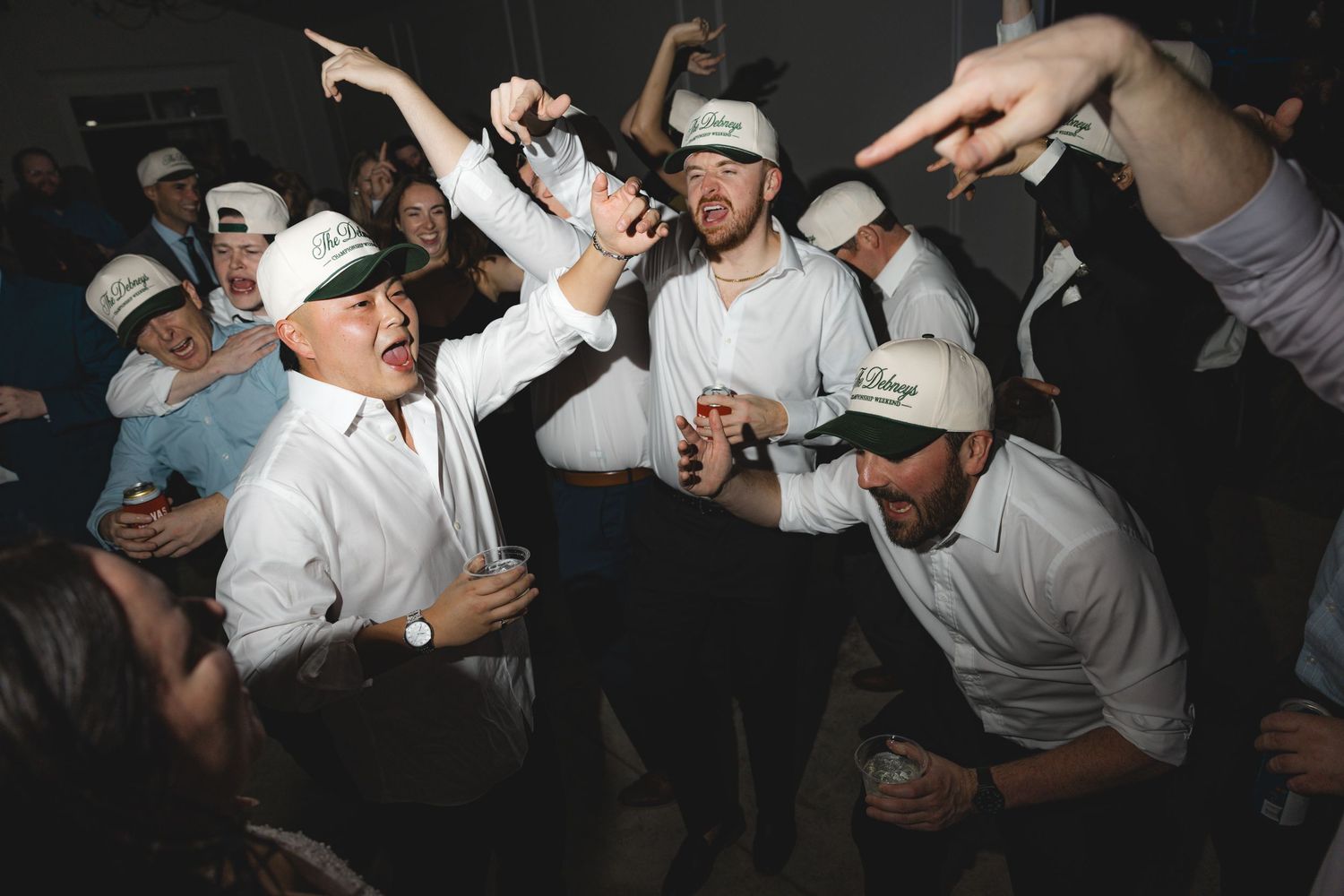 Group of men in white shirts and green caps cheer, point and raise drinks while celebrating at a lively indoor party.