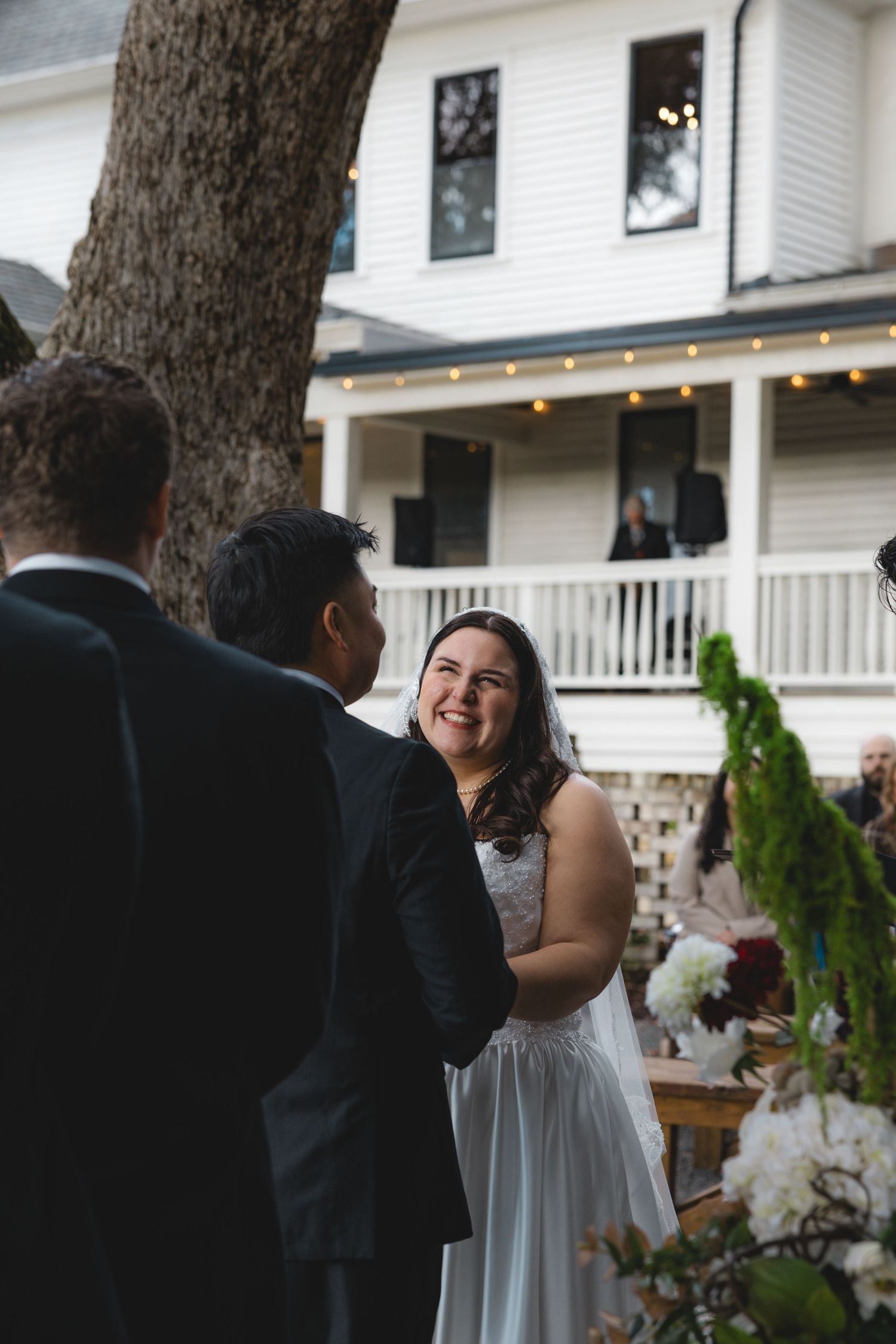 Smiling bride in a white gown and veil exchanges vows with her groom during an outdoor wedding ceremony in front of a white house.