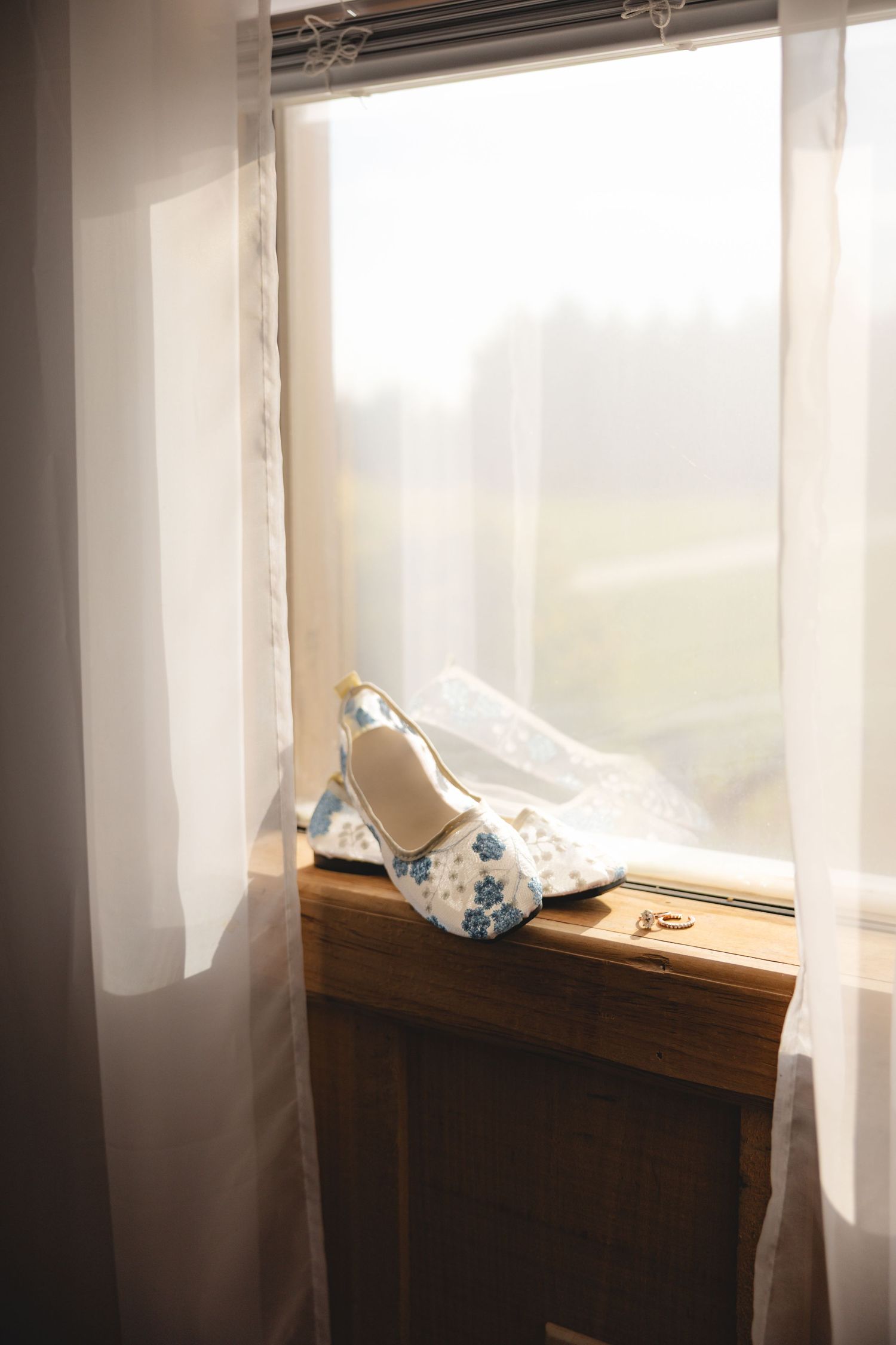White floral wedding shoes with blue flowers resting on wooden windowsill beside gold rings and lace veil.
