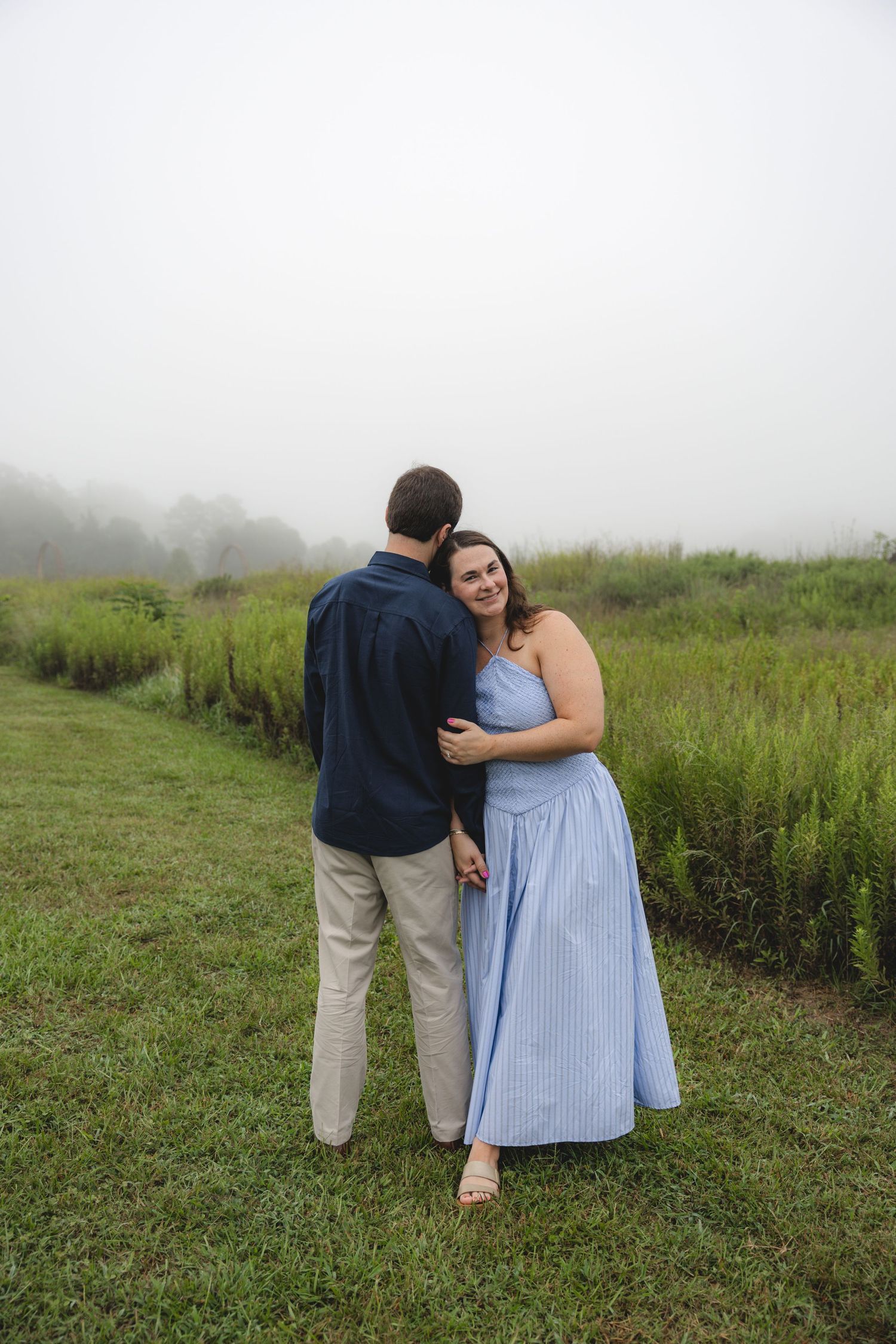 A couple embraces in a misty green field, woman in blue striped dress smiling at camera, man in navy shirt and khakis.