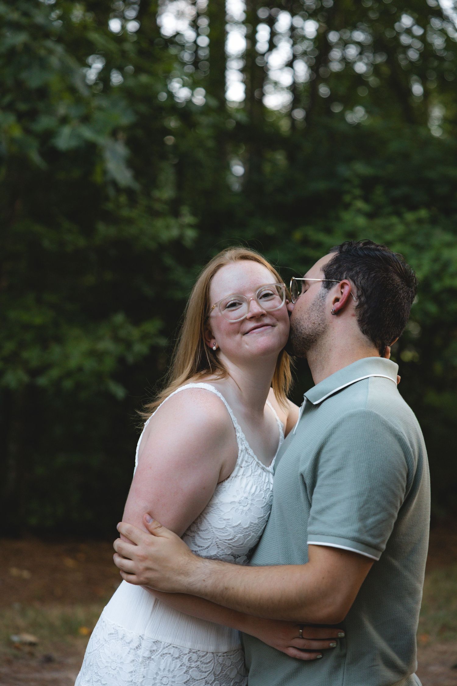 A couple embraces outdoors, man kissing woman's cheek, both wearing glasses, green trees in background.