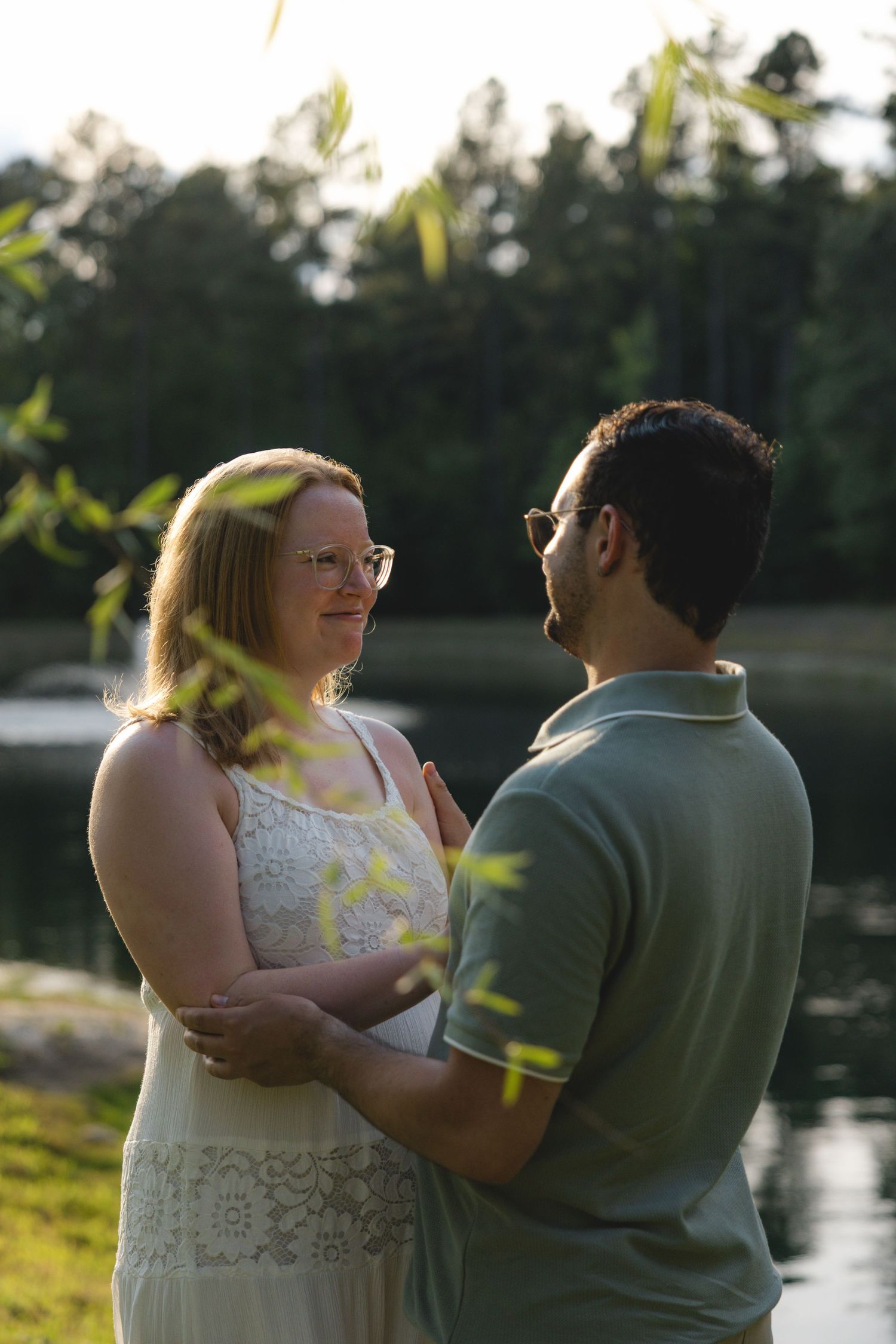 Couple embracing by a sunlit lake, woman in white lace dress and man in green polo, both wearing glasses.