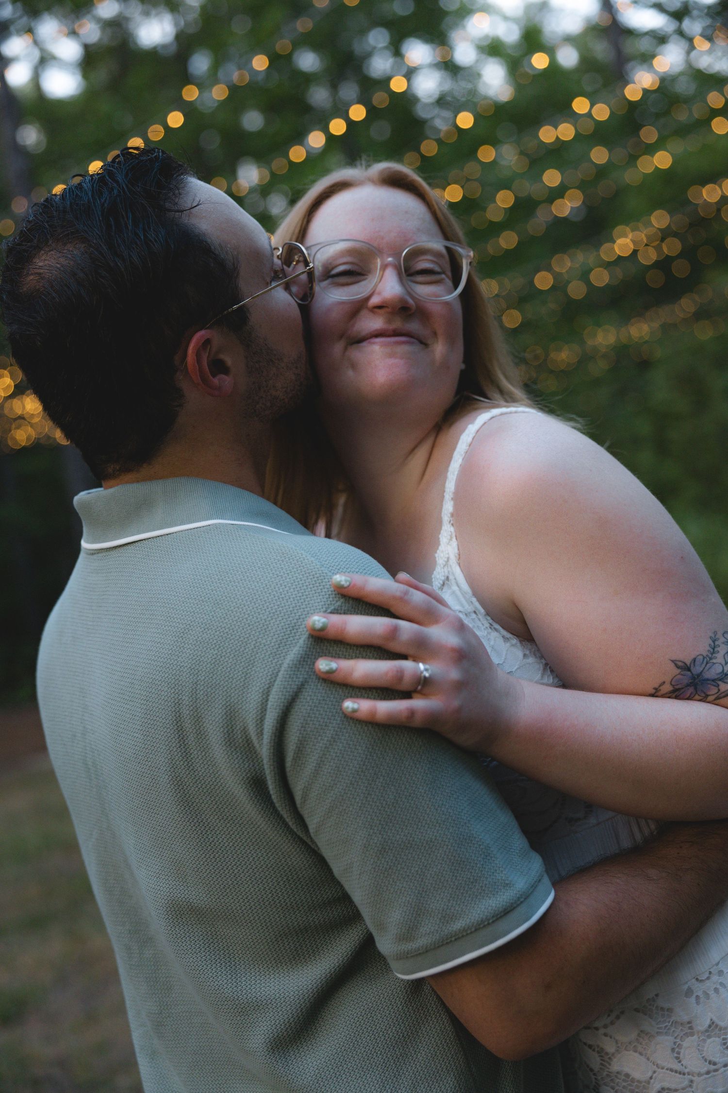 A couple shares a romantic moment outdoors with bokeh string lights, as a man kisses a smiling redhead woman wearing glasses.