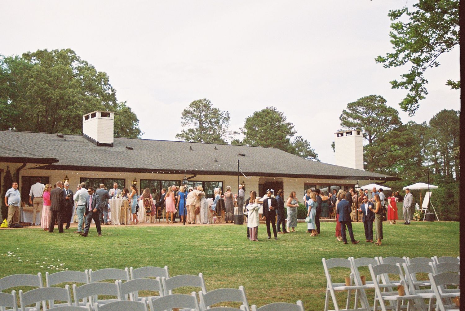 Outdoor wedding reception on a green lawn with guests mingling near a rustic venue surrounded by tall trees.