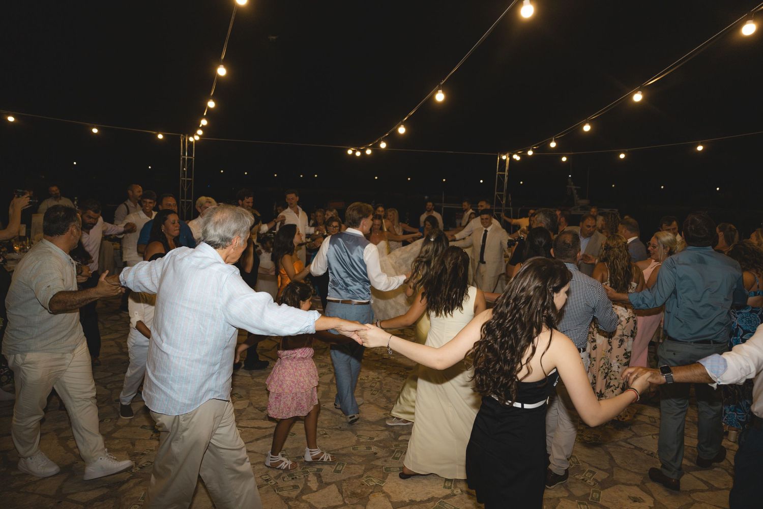 Guests dancing in a circle at an outdoor evening wedding reception under warm string lights.