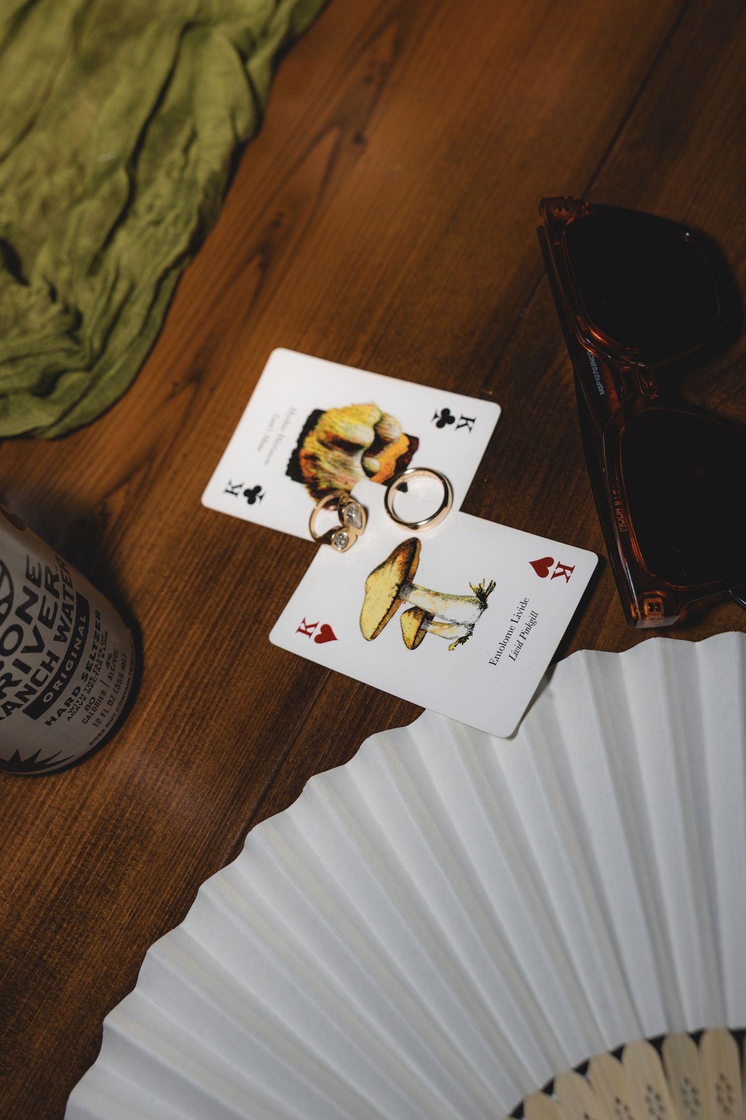 Two mushroom playing cards with a keyring on a wooden table beside sunglasses, a white fan, and a green cloth.