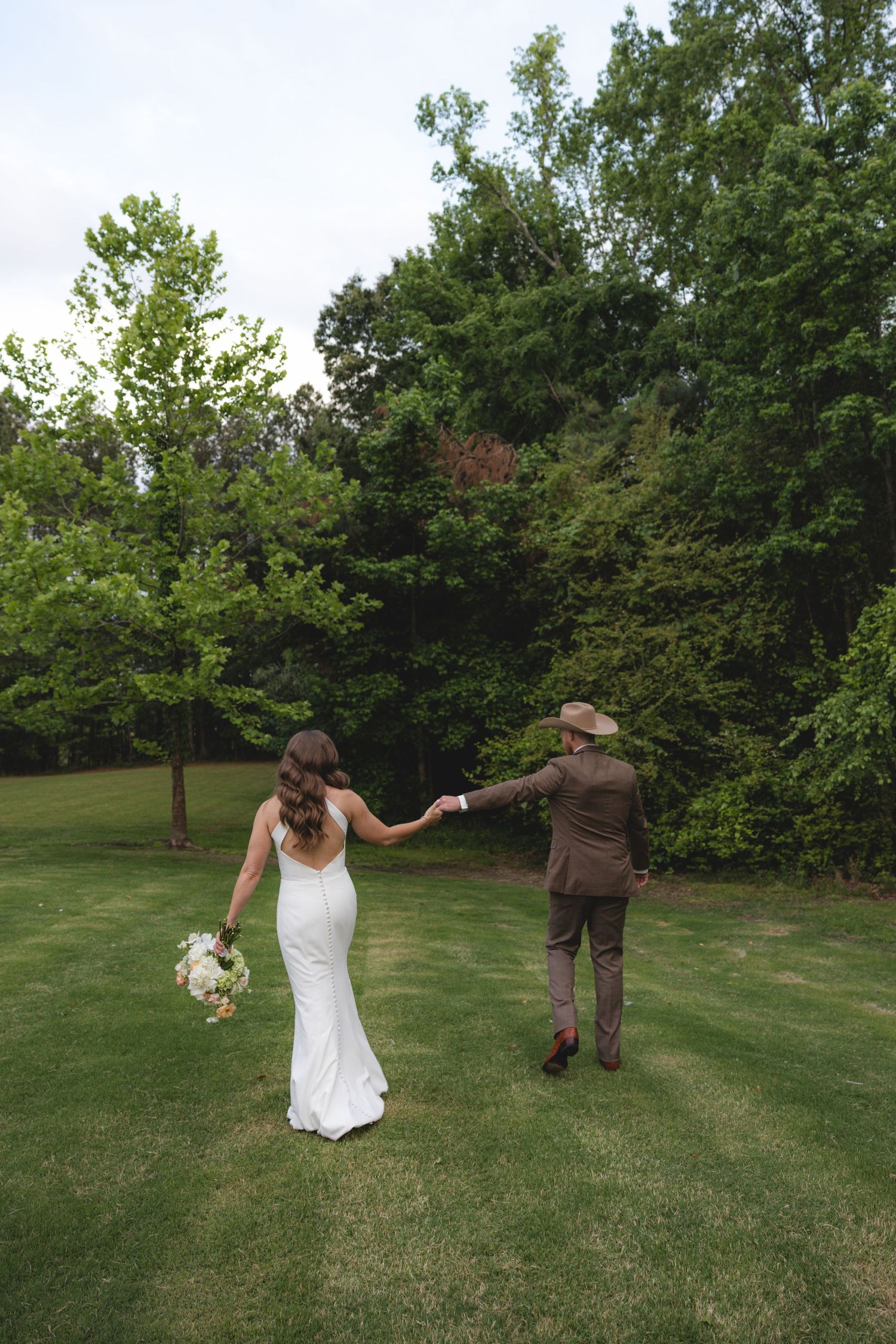 Bride in white gown and groom in brown suit holding hands walking across green lawn toward lush trees.
