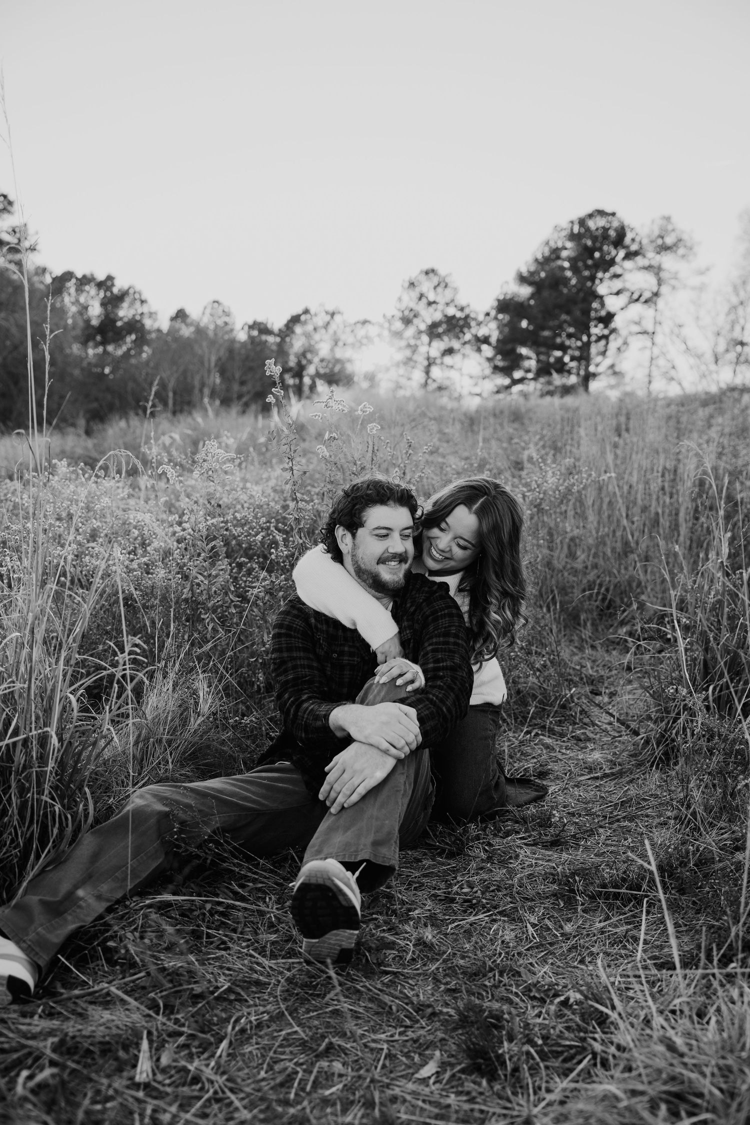 Black and white photo of a couple sitting in a field, smiling and embracing among tall grass with trees behind.