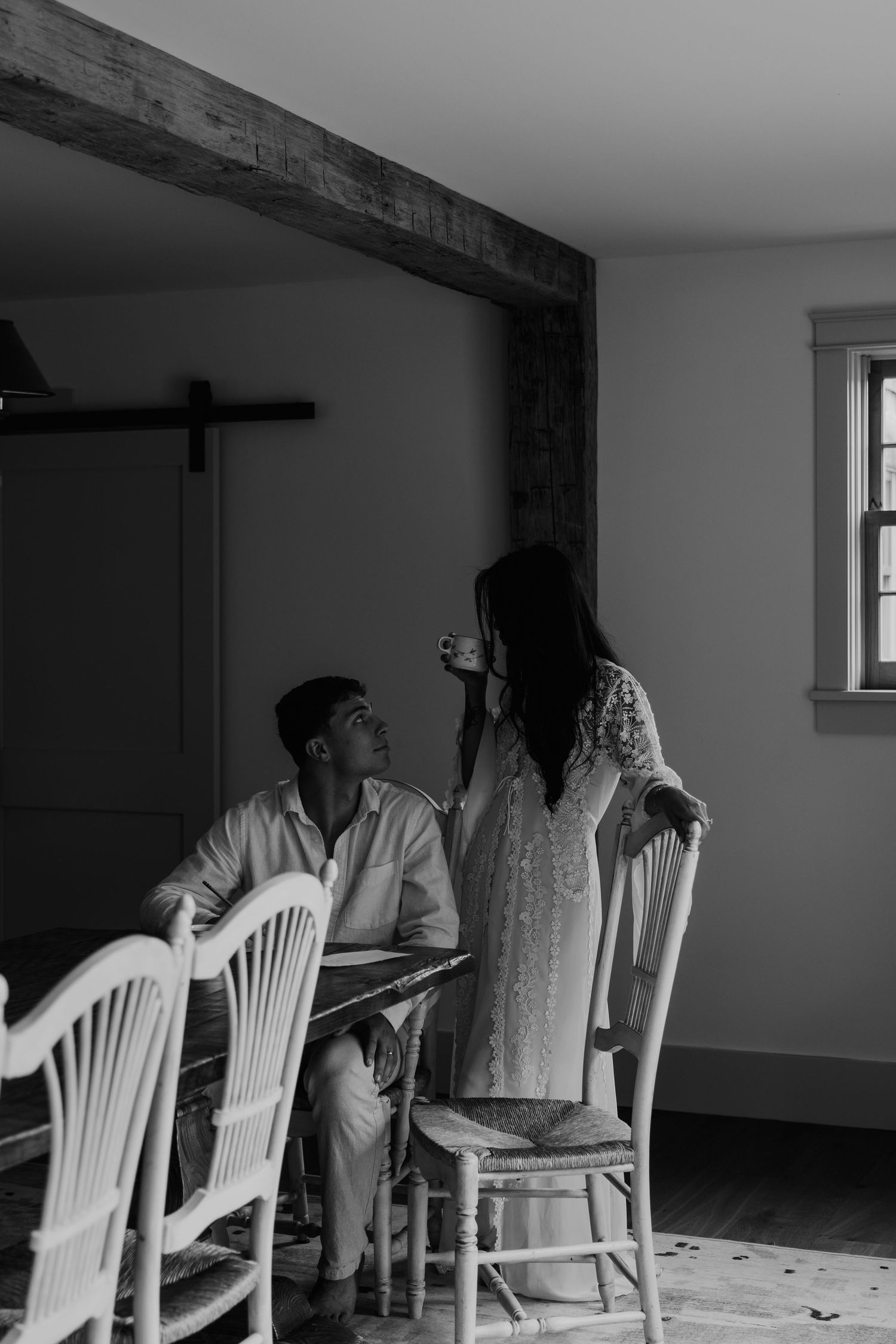 Black and white photo of a couple sharing a tender moment at a rustic dining table in a charming farmhouse interior.