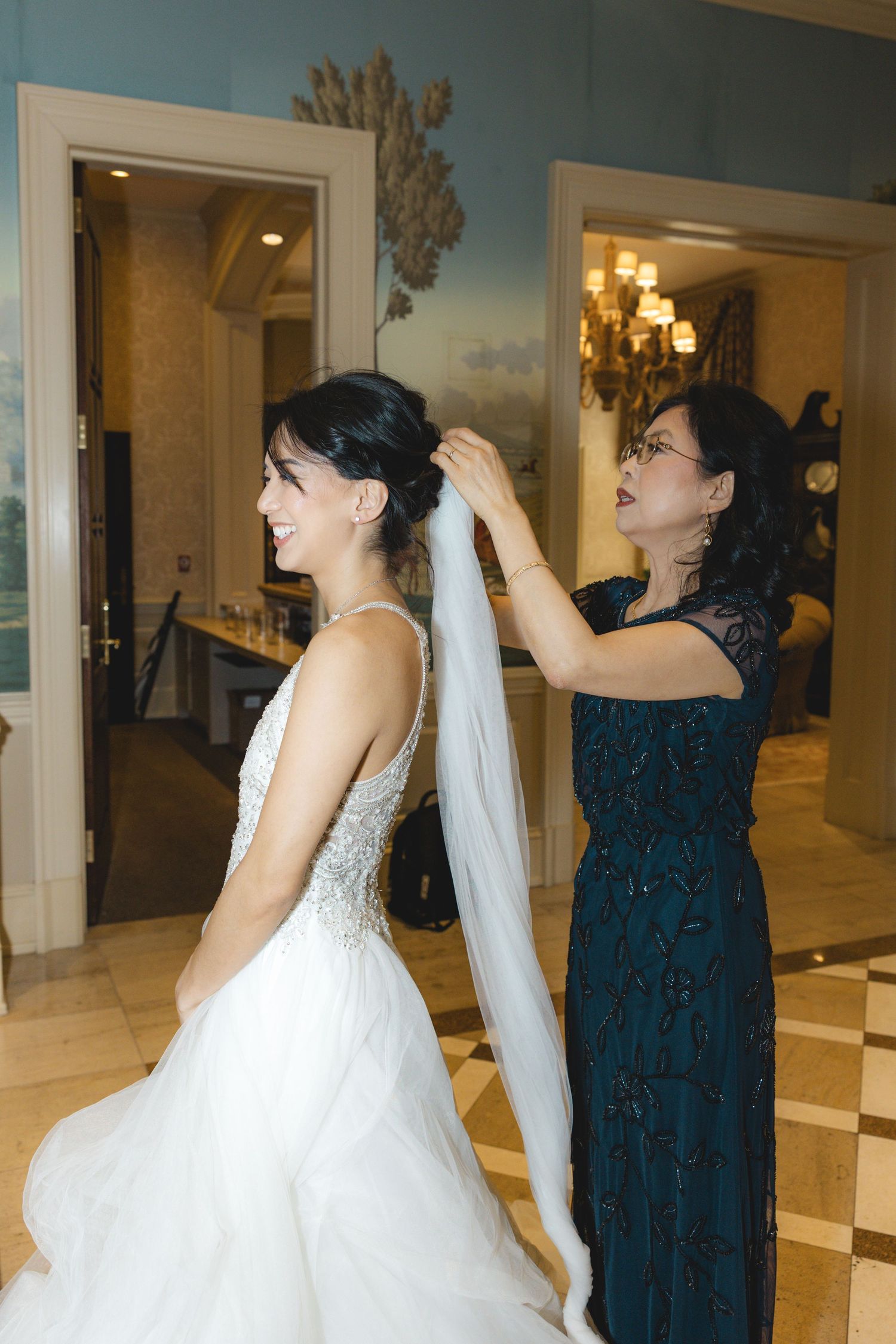 Mother helps smiling bride adjust her long veil in a luxurious hotel bridal suite before the wedding ceremony.