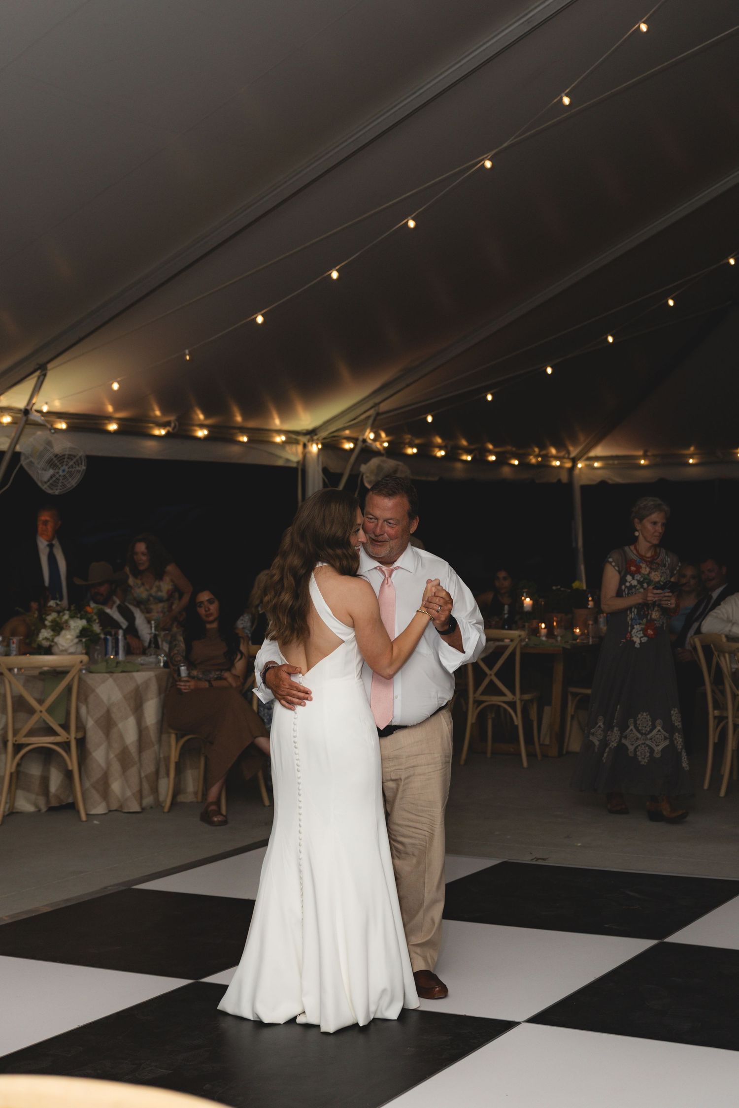 Bride in white gown shares emotional father-daughter dance on checkered floor at elegant tented wedding reception.