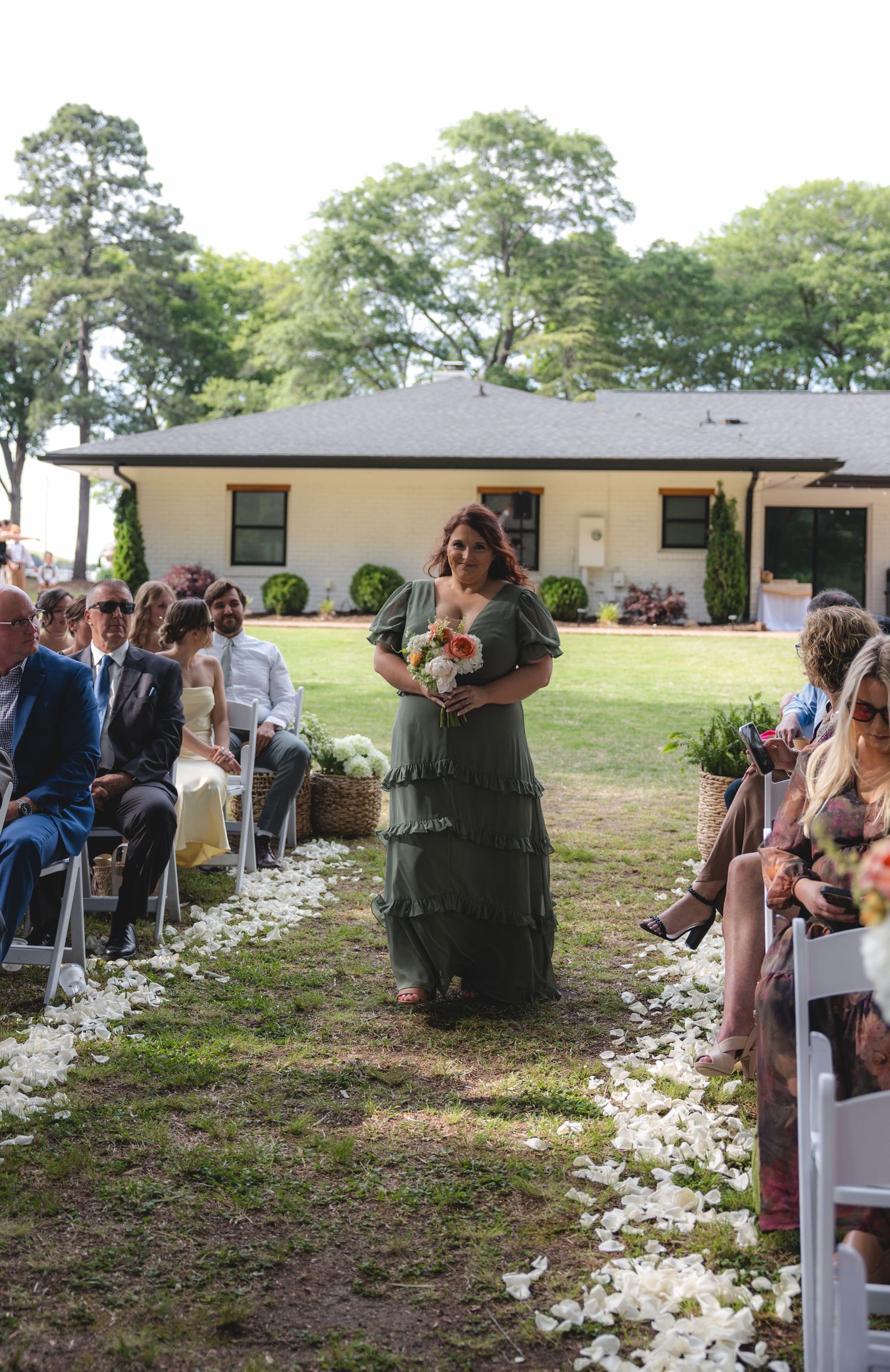 Bridesmaid in sage green ruffled dress carrying peach bouquet walking down outdoor wedding aisle.